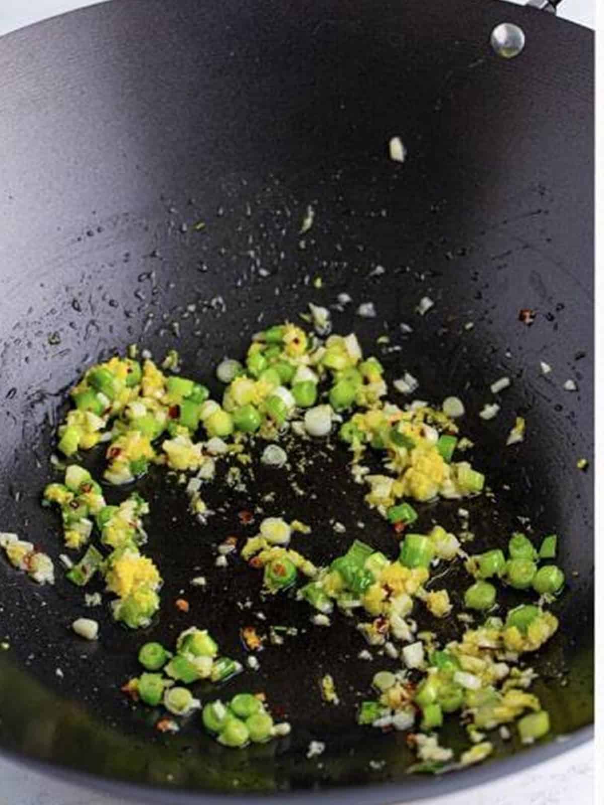Sauteing green onion, garlic, ginger, and chili flakes in a wok until fragrant.