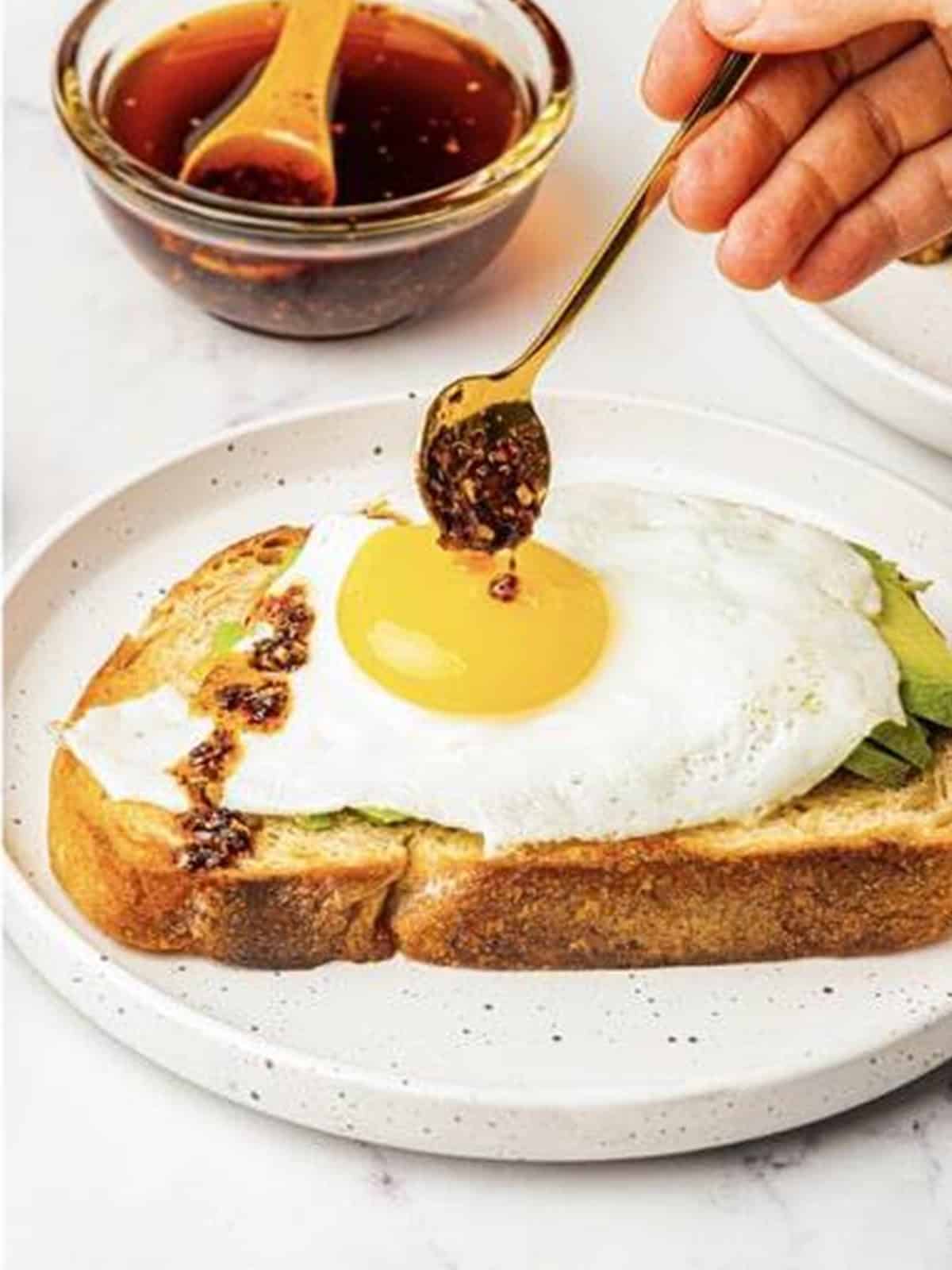 Chili oil being drizzled over a finished avocado toast with a fried egg.