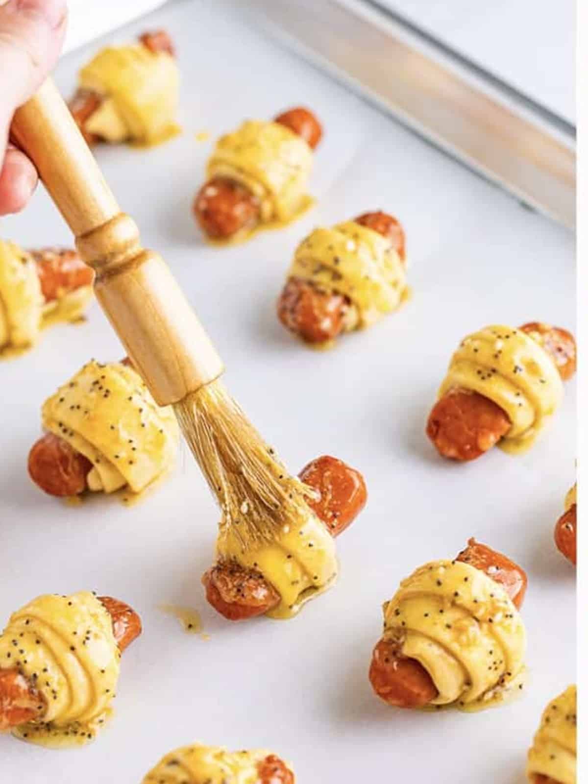 Brushing savory glaze over wrapped pigs in a blanket on a baking sheet.