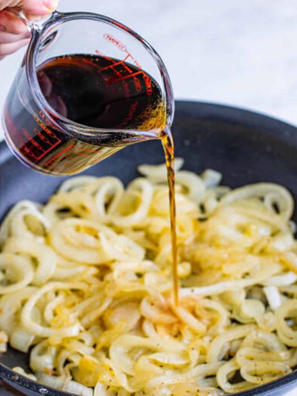 Coca-Cola being poured over softened onions in a skillet.