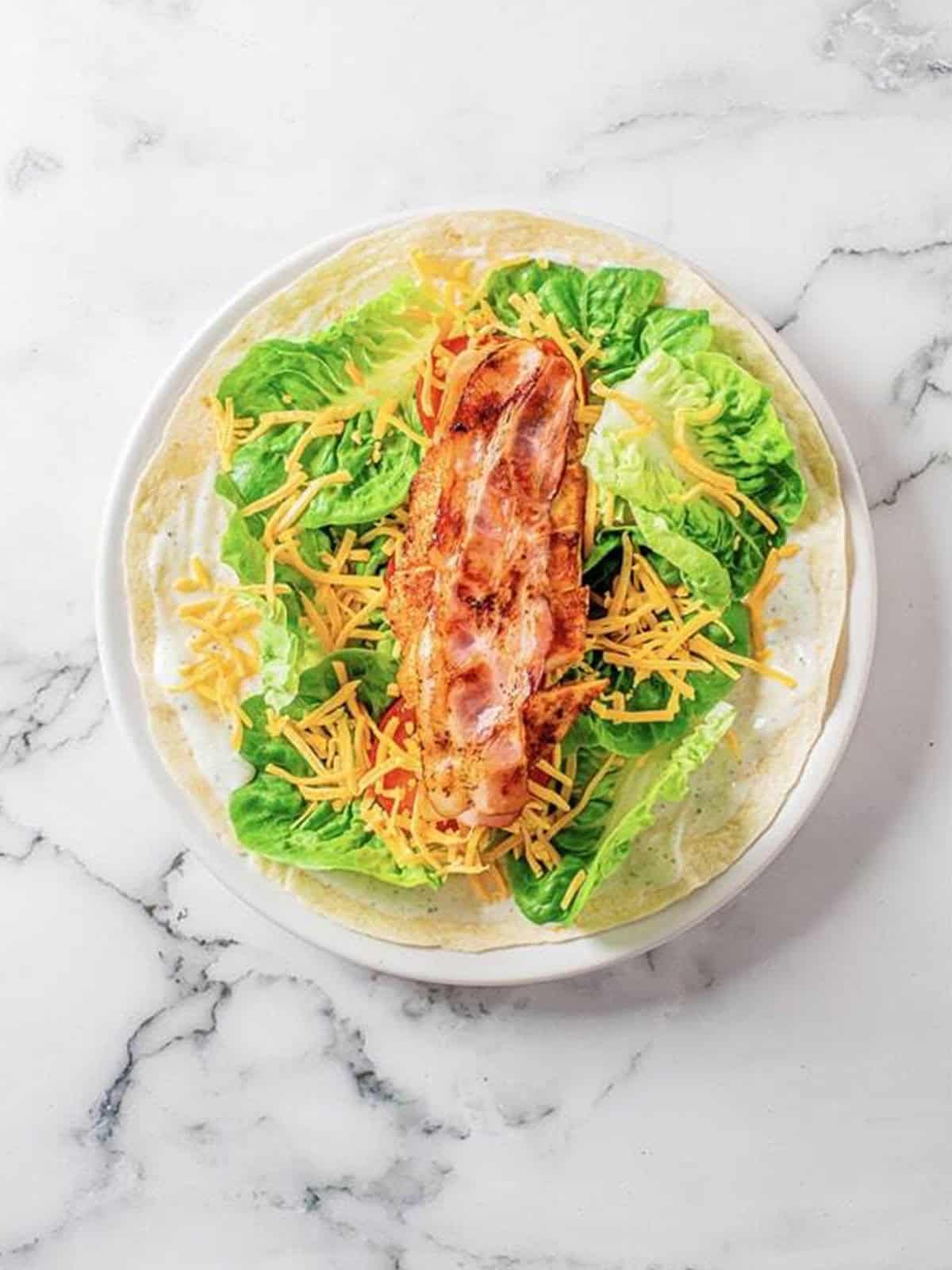 Tomato, lettuce, cheddar, bacon, and chicken layered down the center of a tortilla.