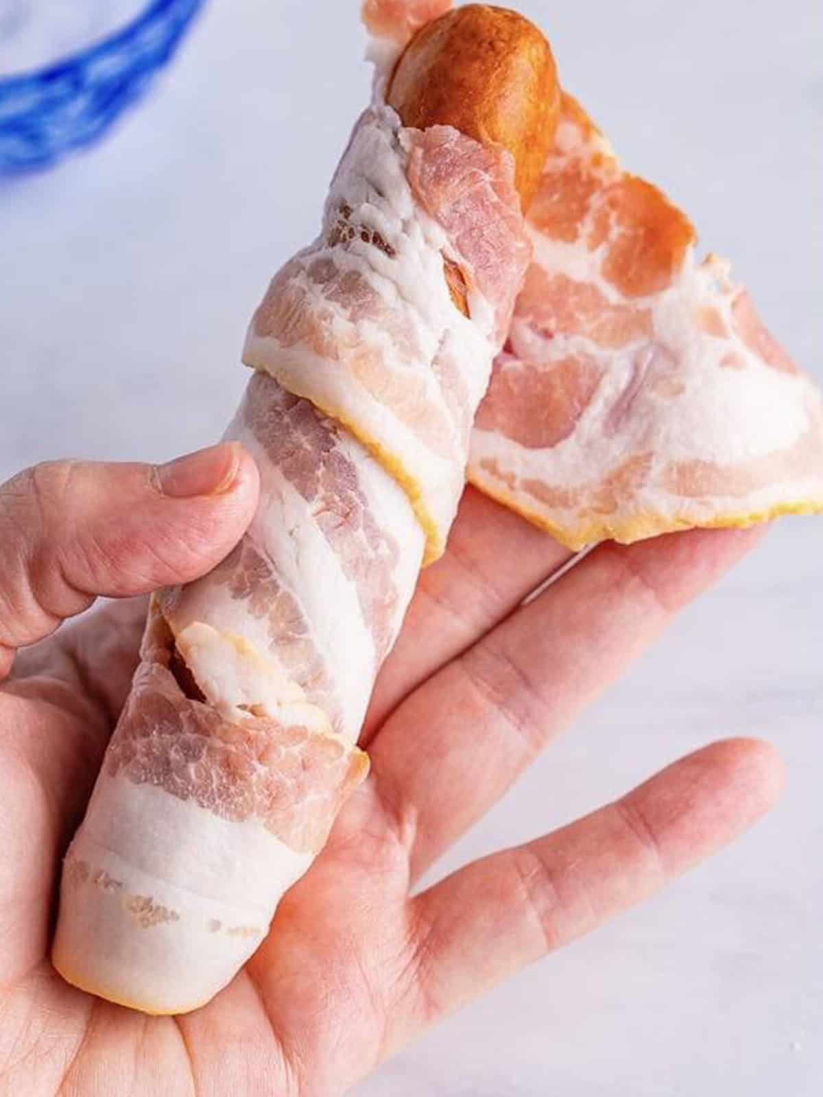 A bratwurst being wrapped in a strip of raw bacon.