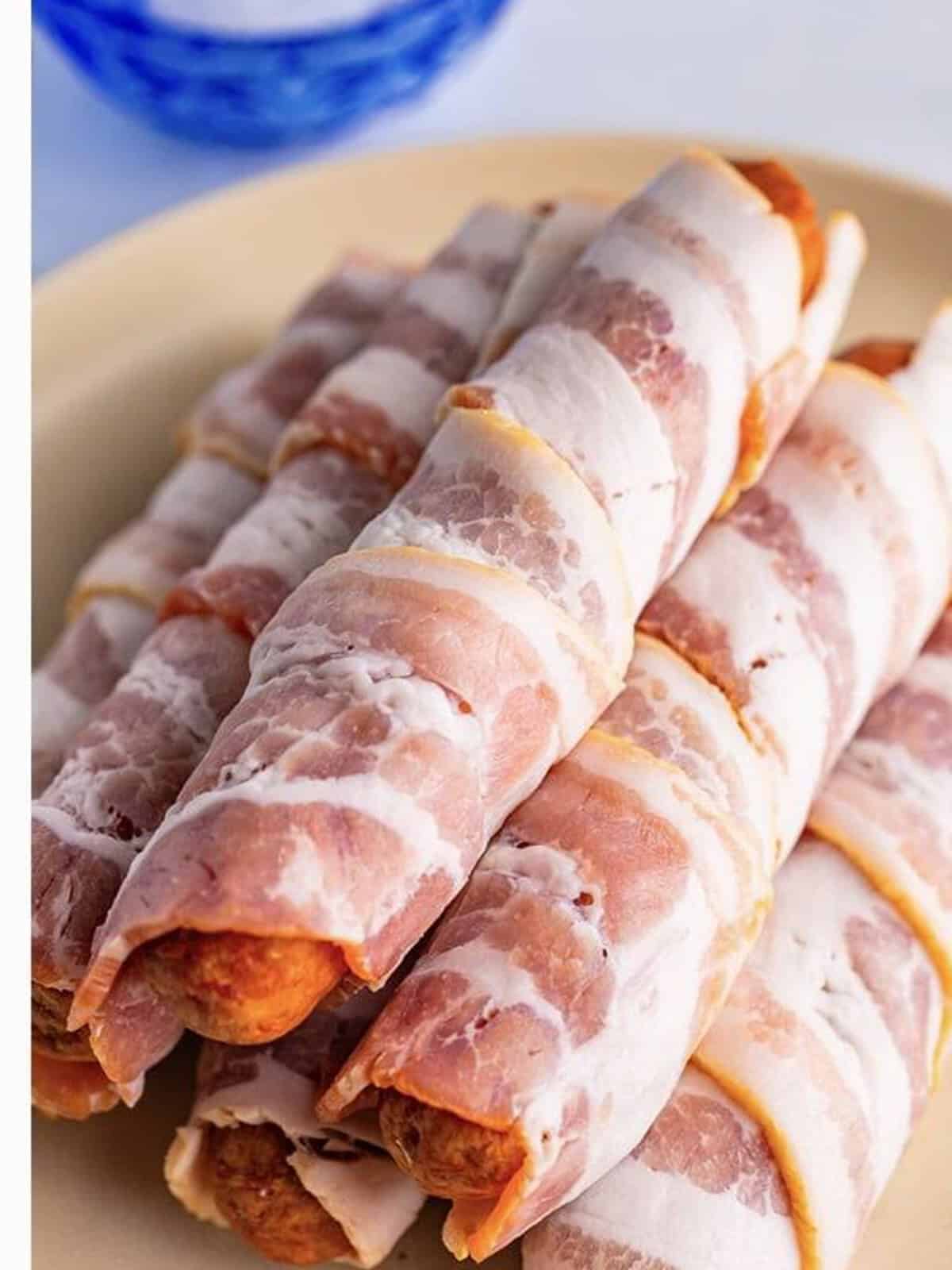 Several bacon wrapped bratwursts arranged on a plate before cooking.