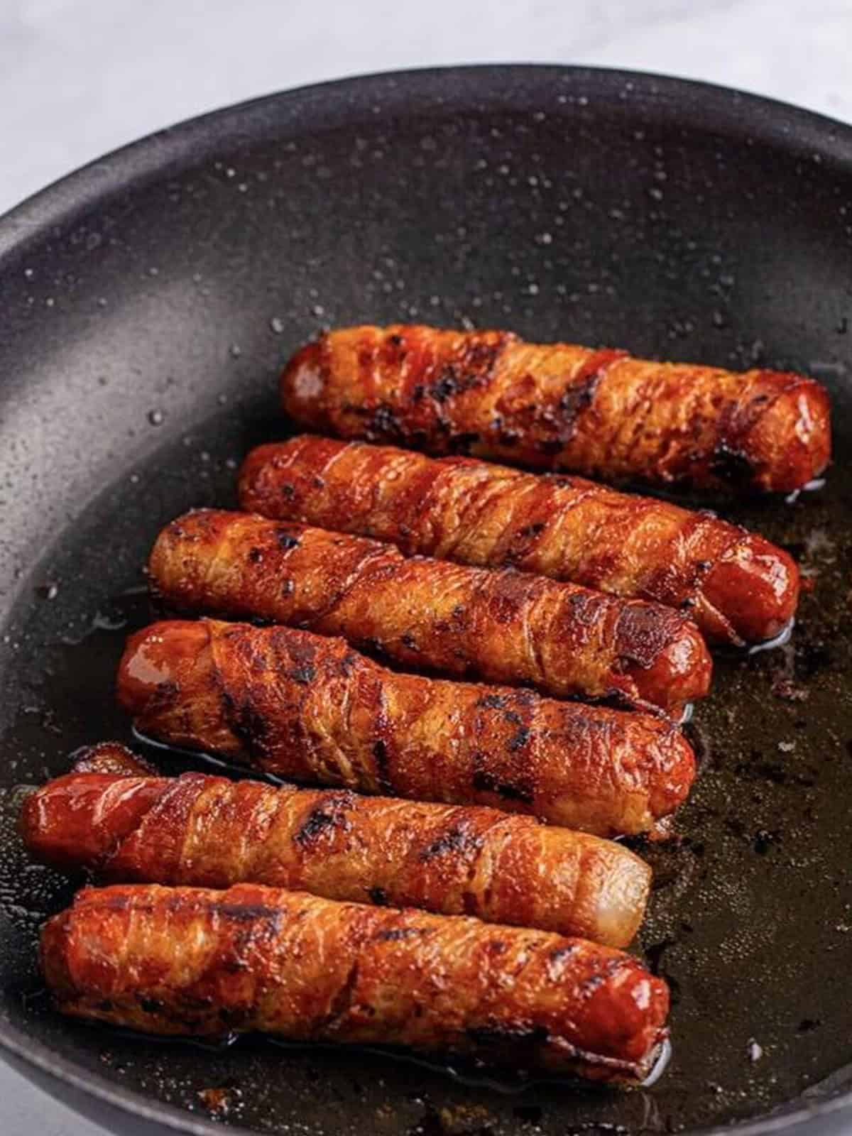 Bacon wrapped bratwursts cooking and browning in a skillet.