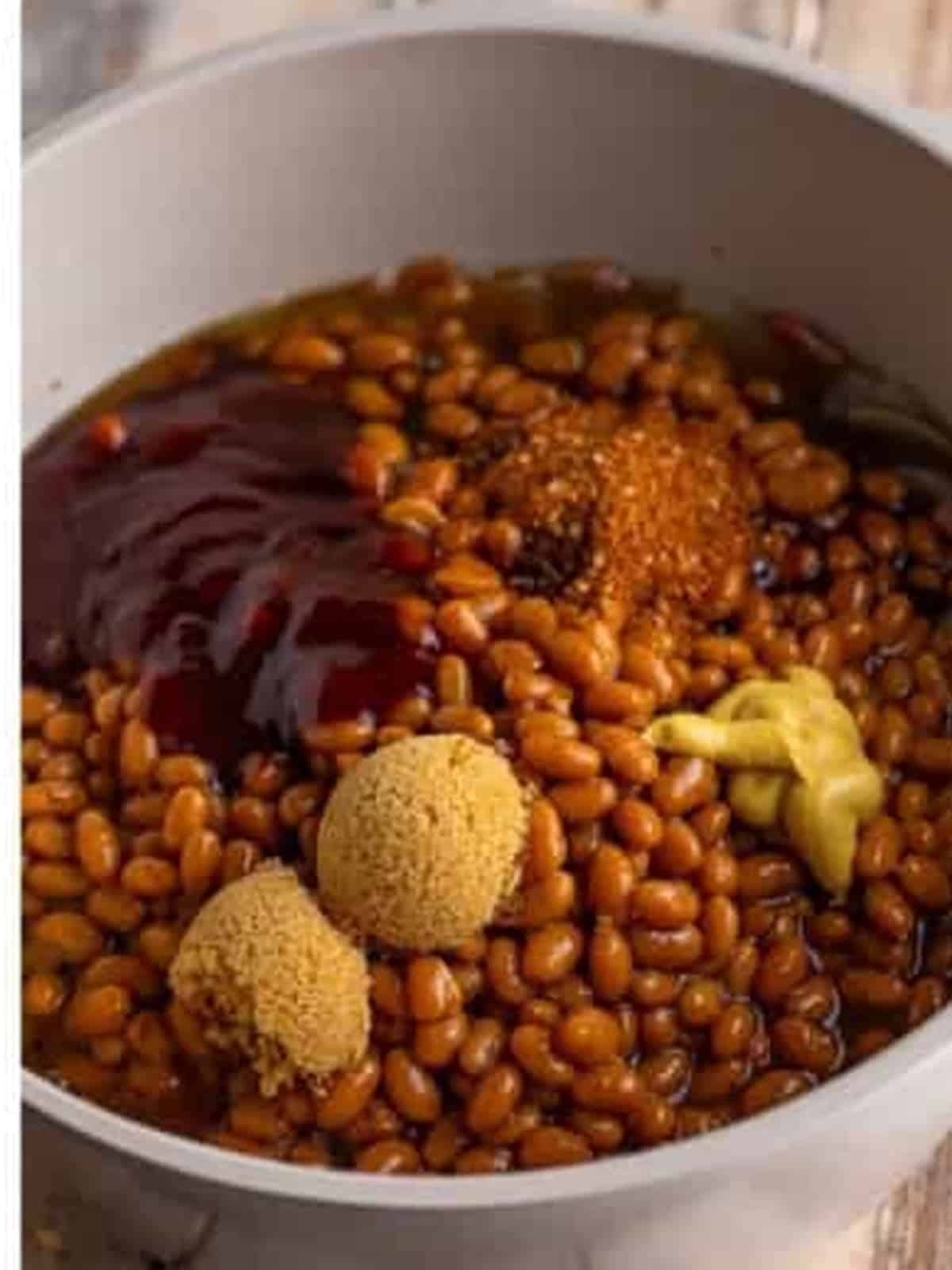 Canned baked beans topped with BBQ sauce, brown sugar, mustard, and seasonings in a pot.