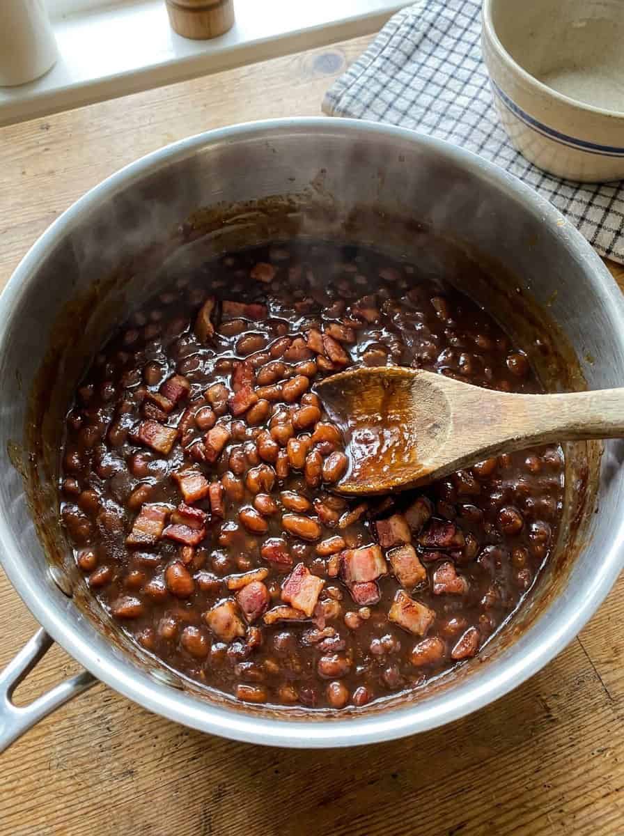 Baked beans simmering thick and syrupy with crispy bacon in a saucepan.