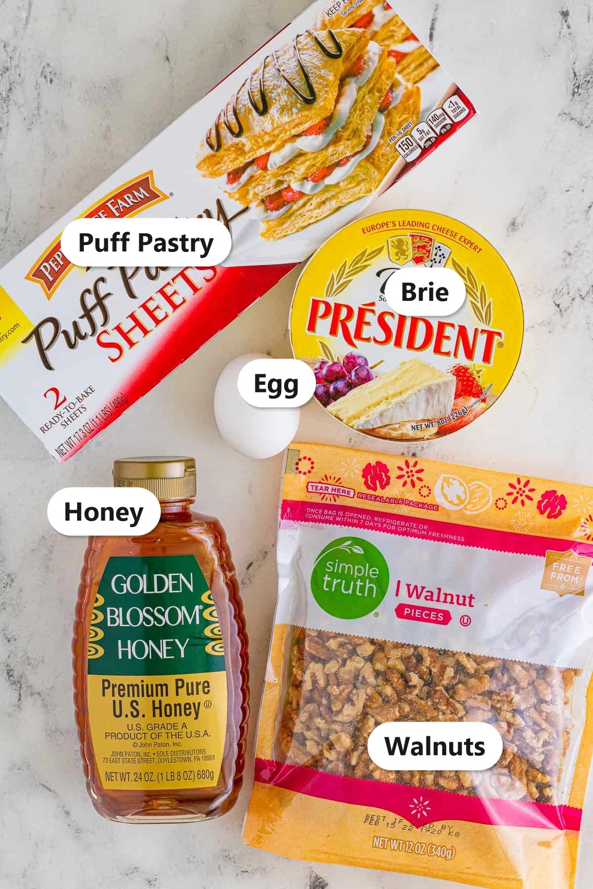 Labeled ingredients for baked brie in puff pastry including puff pastry sheets, a wheel of brie, honey, walnuts, and an egg.