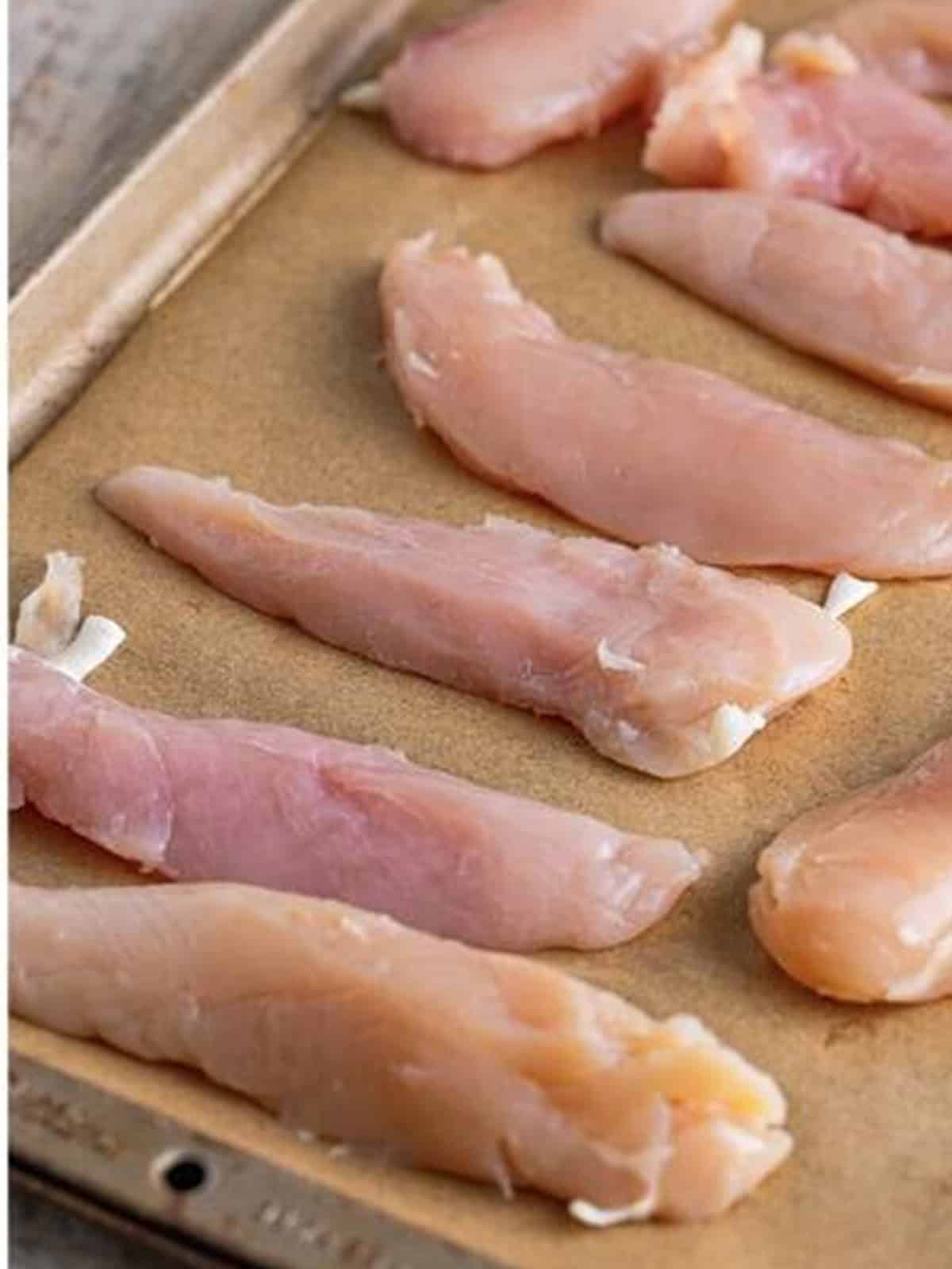 Raw chicken tenderloins arranged on a parchment lined sheet tray for baked chicken tenders.