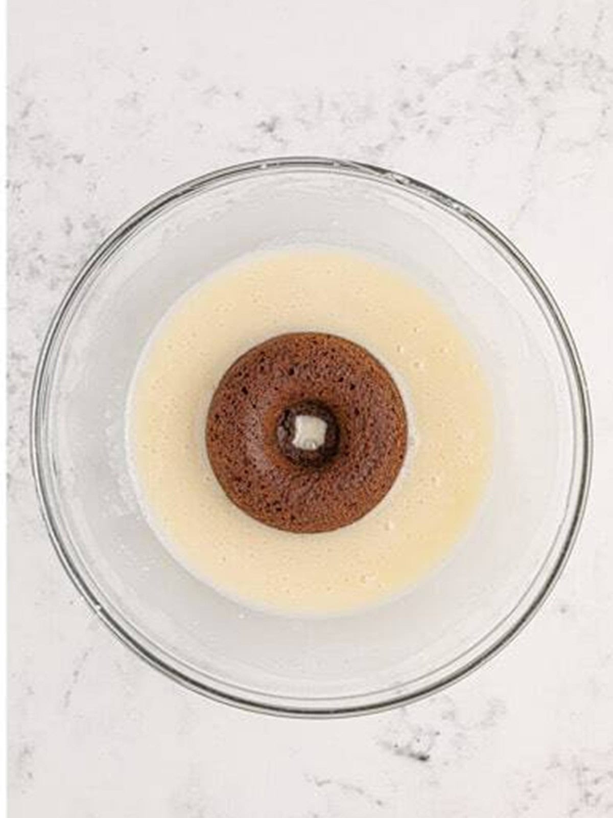 A baked chocolate donut being dipped into a simple vanilla glaze.
