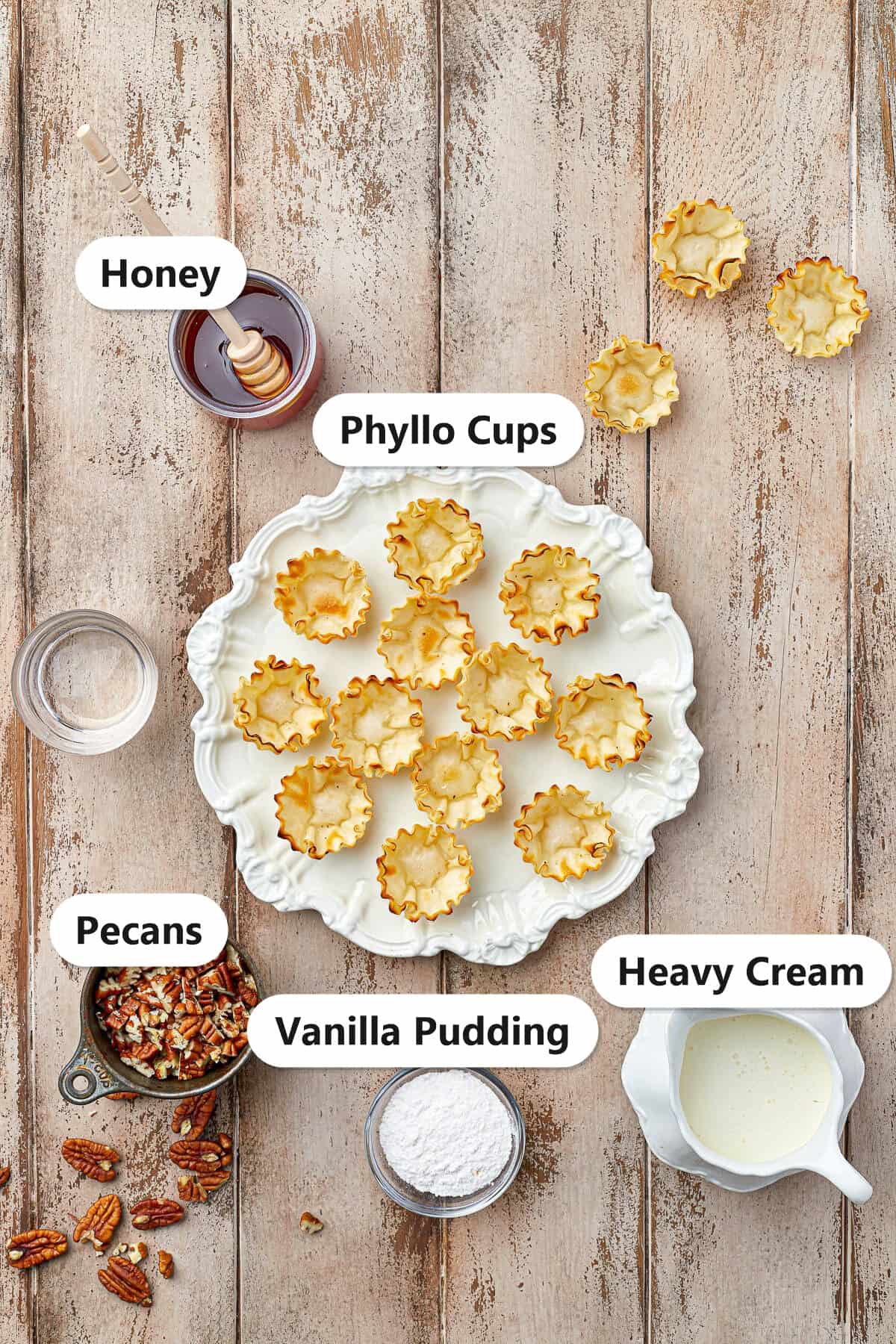 Baklava Bites ingredients including phyllo cups, honey, pecans, vanilla pudding, and heavy cream labeled on a wood counter.