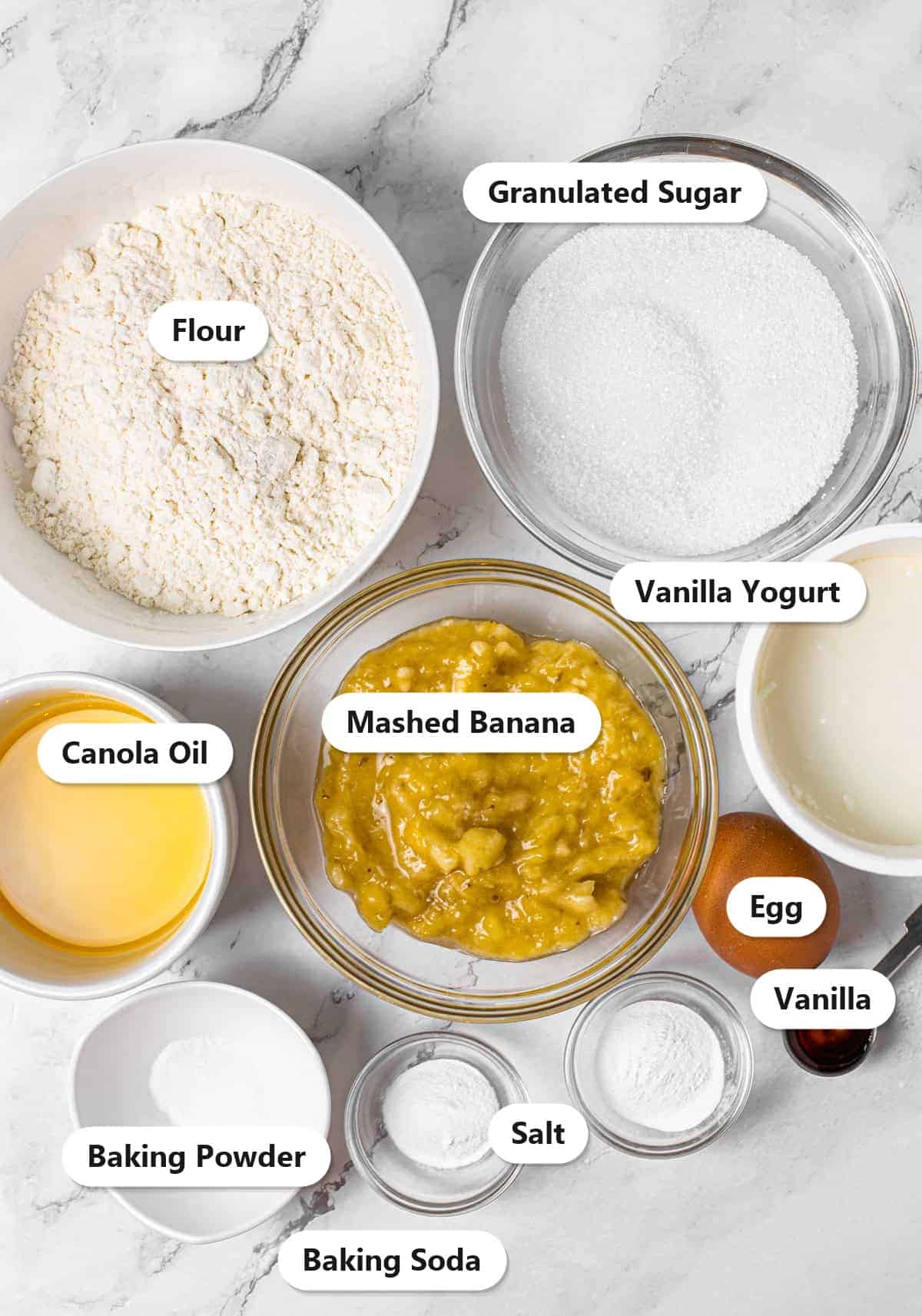 Ingredients to make banana cake including flour, granulated sugar, mashed banana, vanilla yogurt, canola oil, egg, and baking staples.