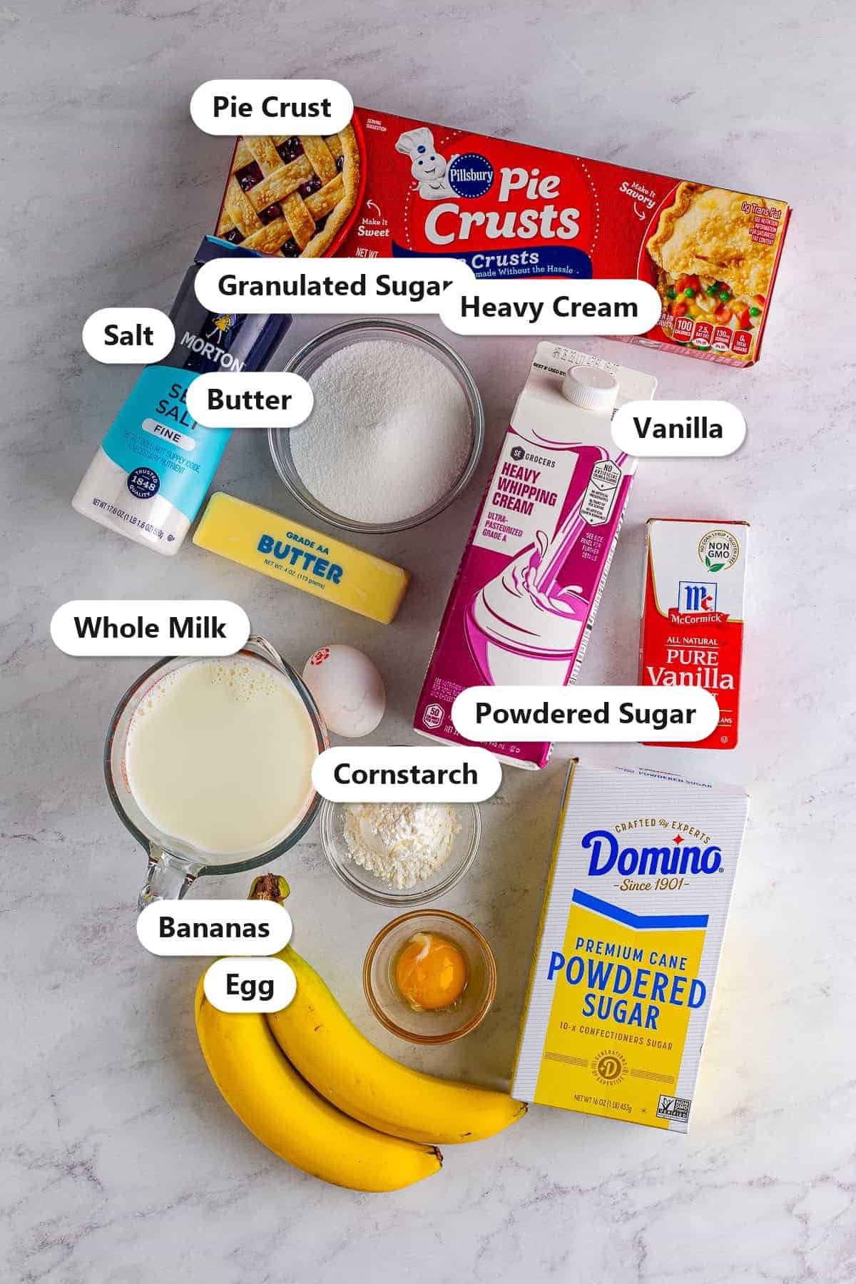 Labeled ingredients to make banana cream pie including pie crust, whole milk, sugar, cornstarch, eggs, butter, bananas, and heavy cream.