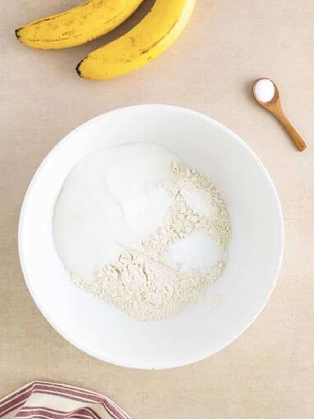 Flour, sugar, baking powder, baking soda, and salt whisked together in a white bowl for banana cupcakes.