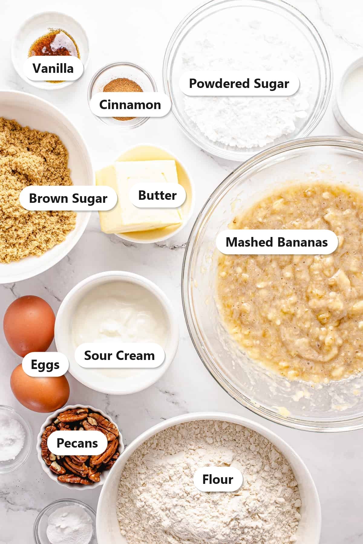 Labeled ingredients for banana nut bread including mashed bananas, flour, brown sugar, butter, eggs, sour cream, pecans, cinnamon, vanilla, and powdered sugar.