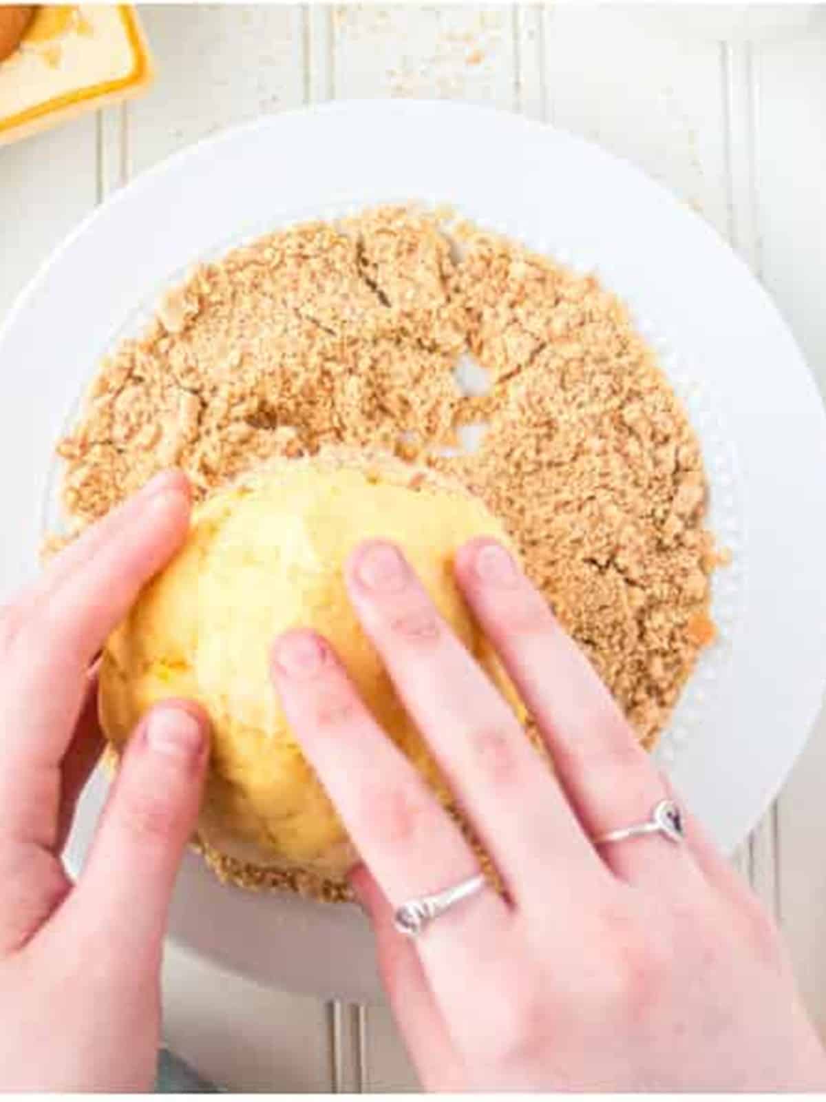 Hands rolling the chilled banana pudding cheese ball in vanilla wafer crumbs.