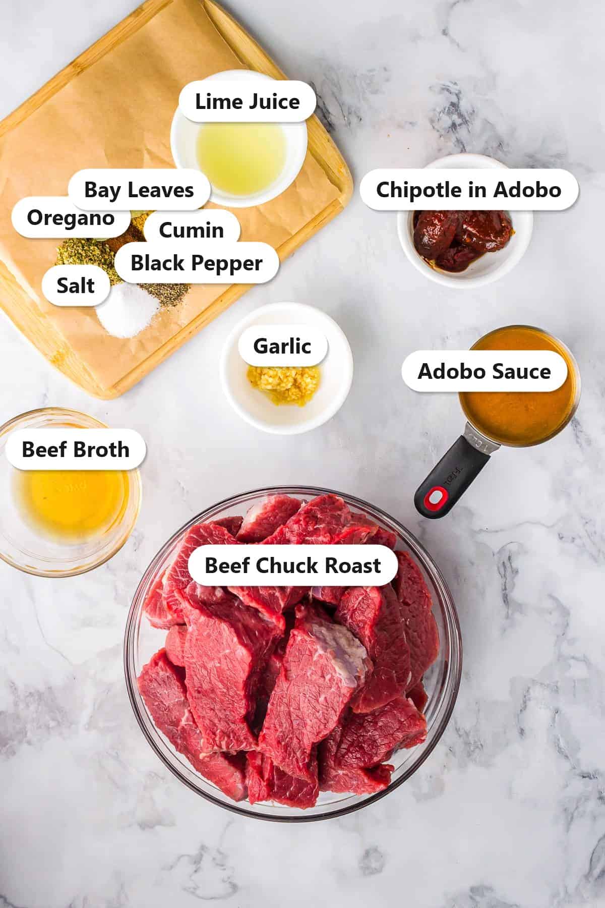 Labeled ingredients for barbacoa including beef chuck roast, beef broth, chipotle in adobo, garlic, cumin, oregano, and lime juice on a marble surface.