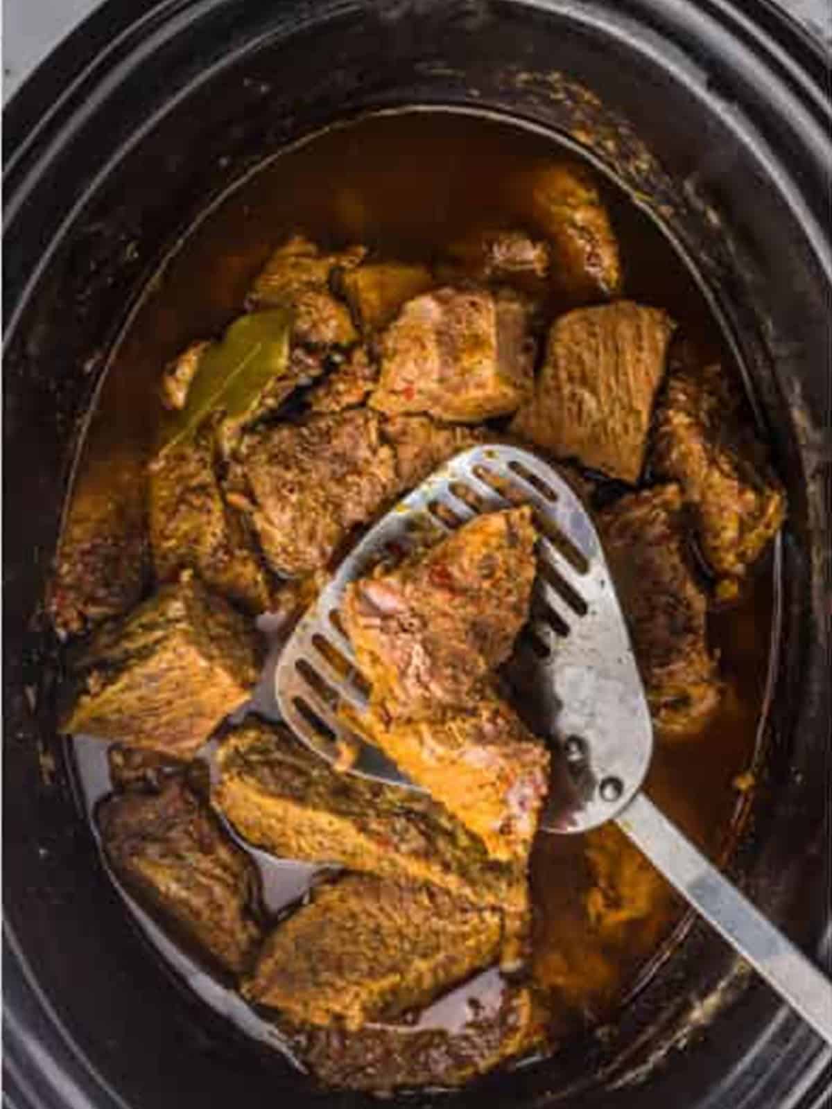 Shredding the cooked beef in the slow cooker with a potato masher for barbacoa.