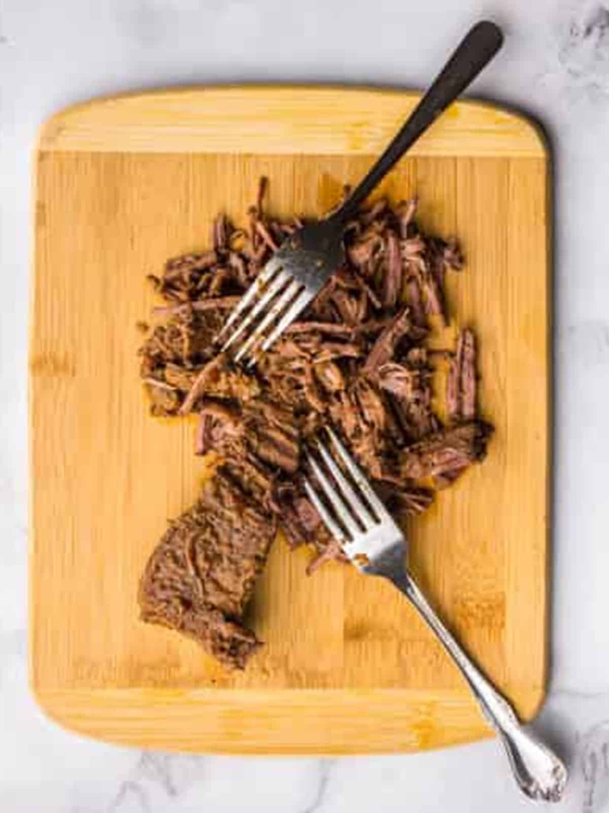 Shredded barbacoa beef on a wooden cutting board pulled apart with two forks.