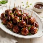 A white platter piled with glossy BBQ bacon wrapped meatballs on toothpicks garnished with sliced scallions.