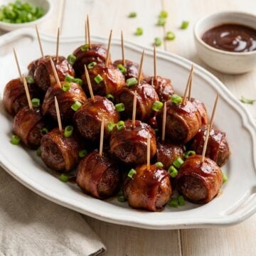A white platter piled with glossy BBQ bacon wrapped meatballs on toothpicks garnished with sliced scallions.