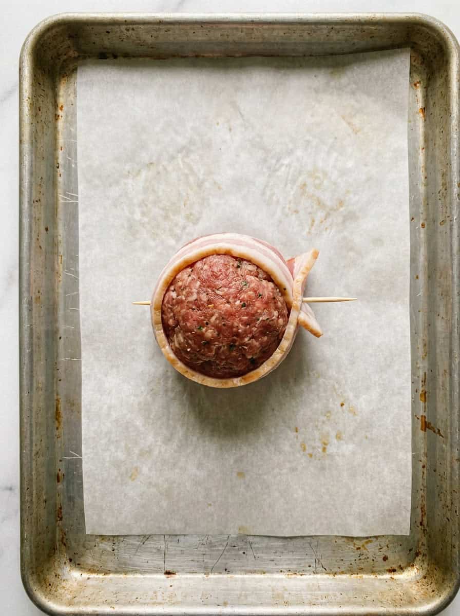 A meatball wrapped in a half slice of bacon and secured with a wooden toothpick on a sheet tray.