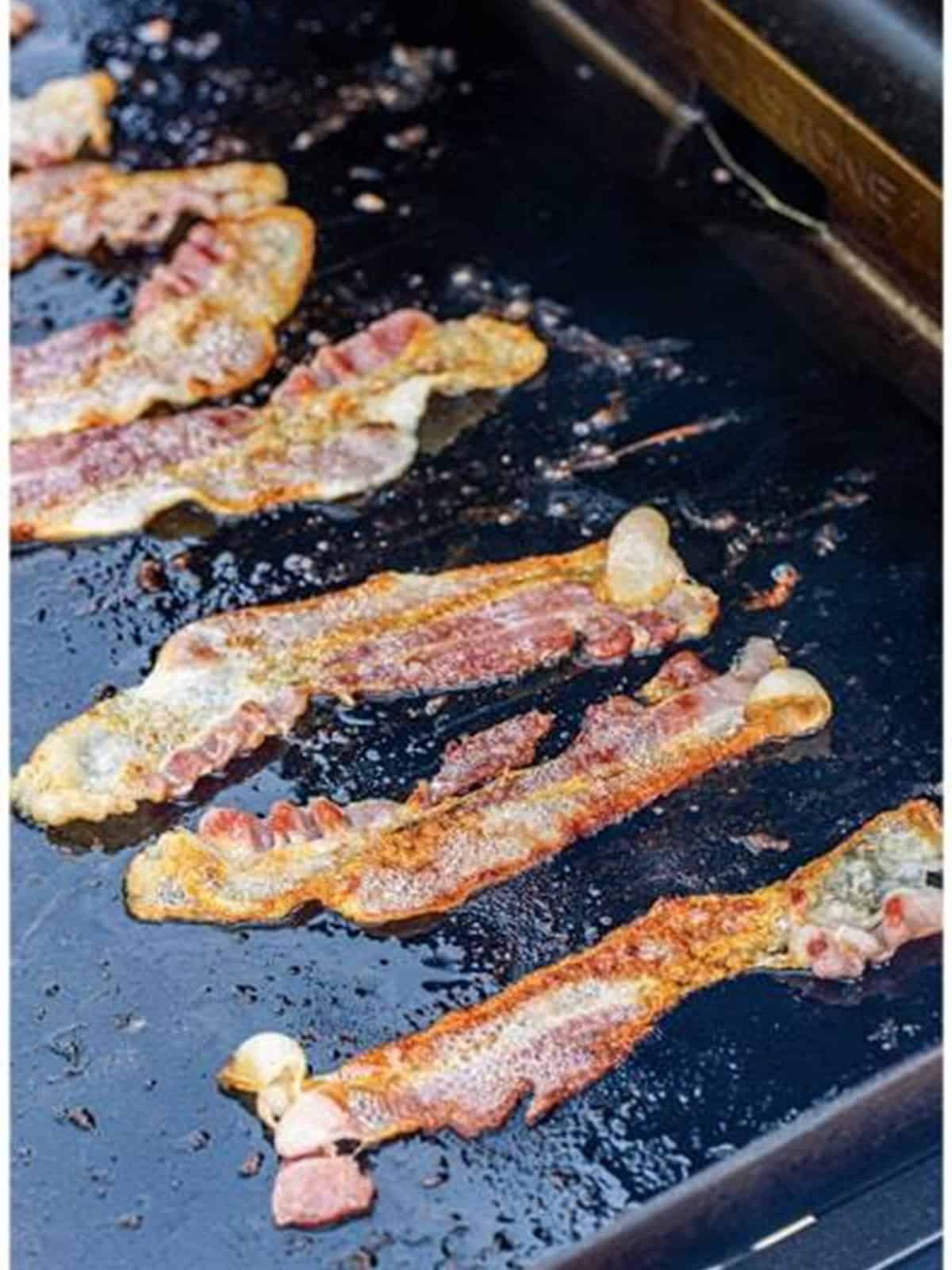 Bacon strips cooking until crispy on a flat top griddle.