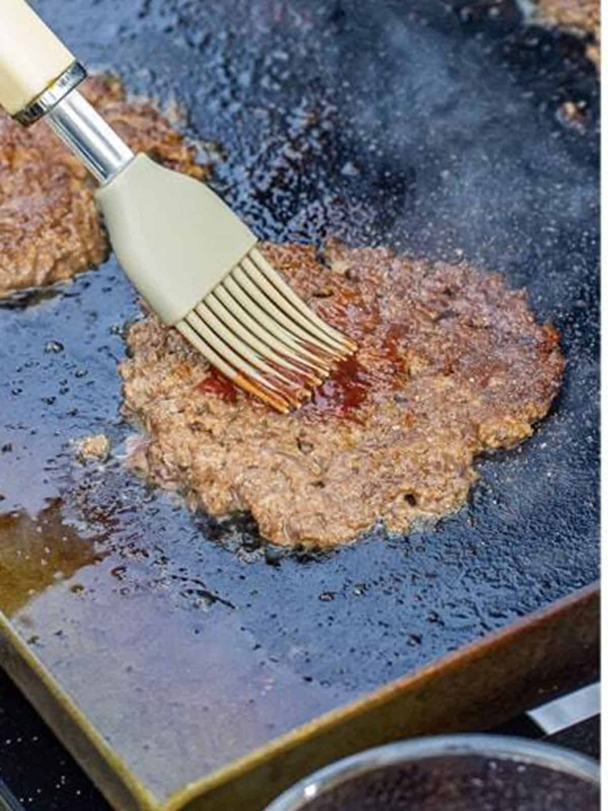 A smashed burger patty being brushed with BBQ sauce.