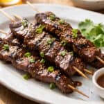 Grilled Asian beef skewers on a platter with char marks, sesame seeds, and green onions, served with a dipping sauce.