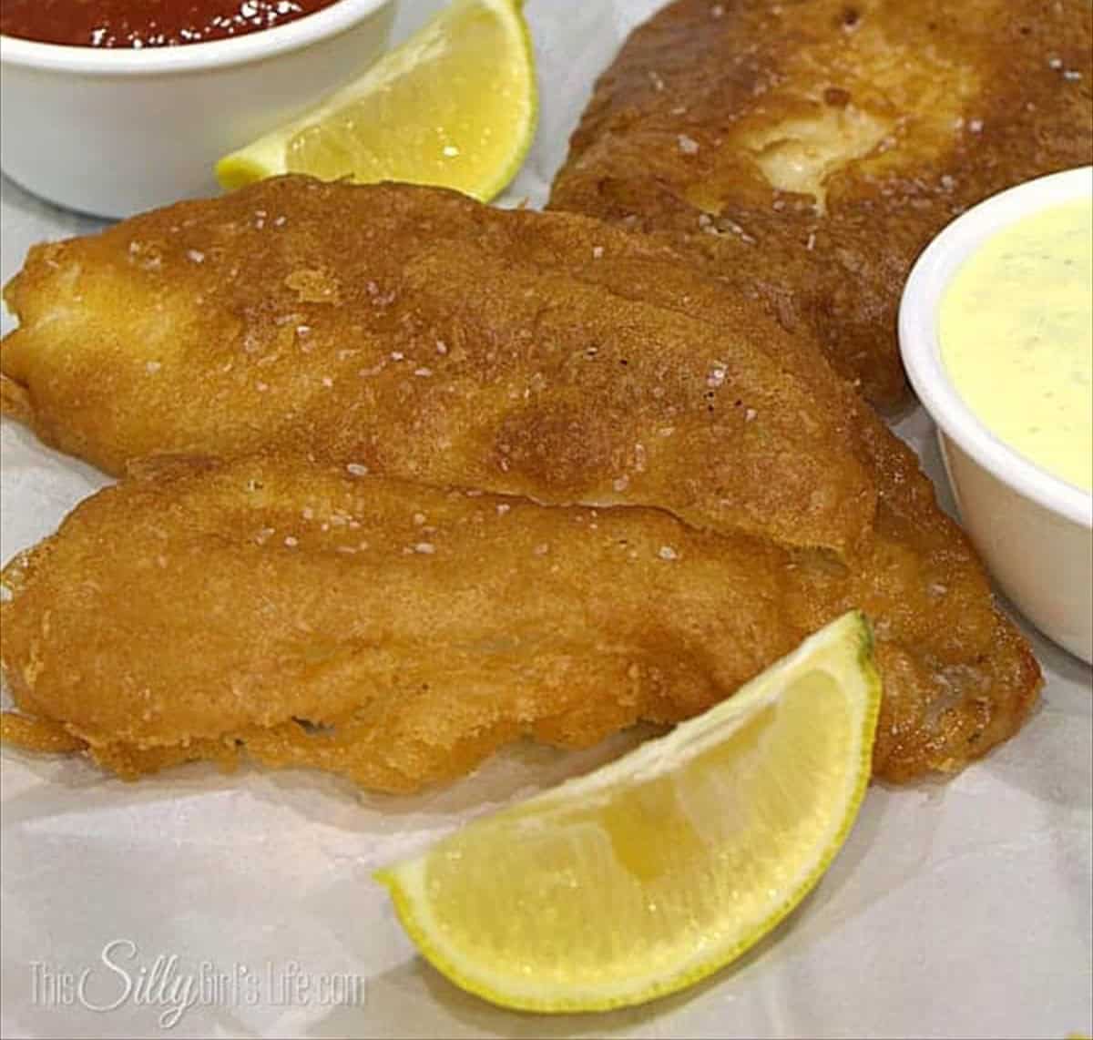 Golden fried beer battered fish fillets sprinkled with salt next to lemon wedges.
