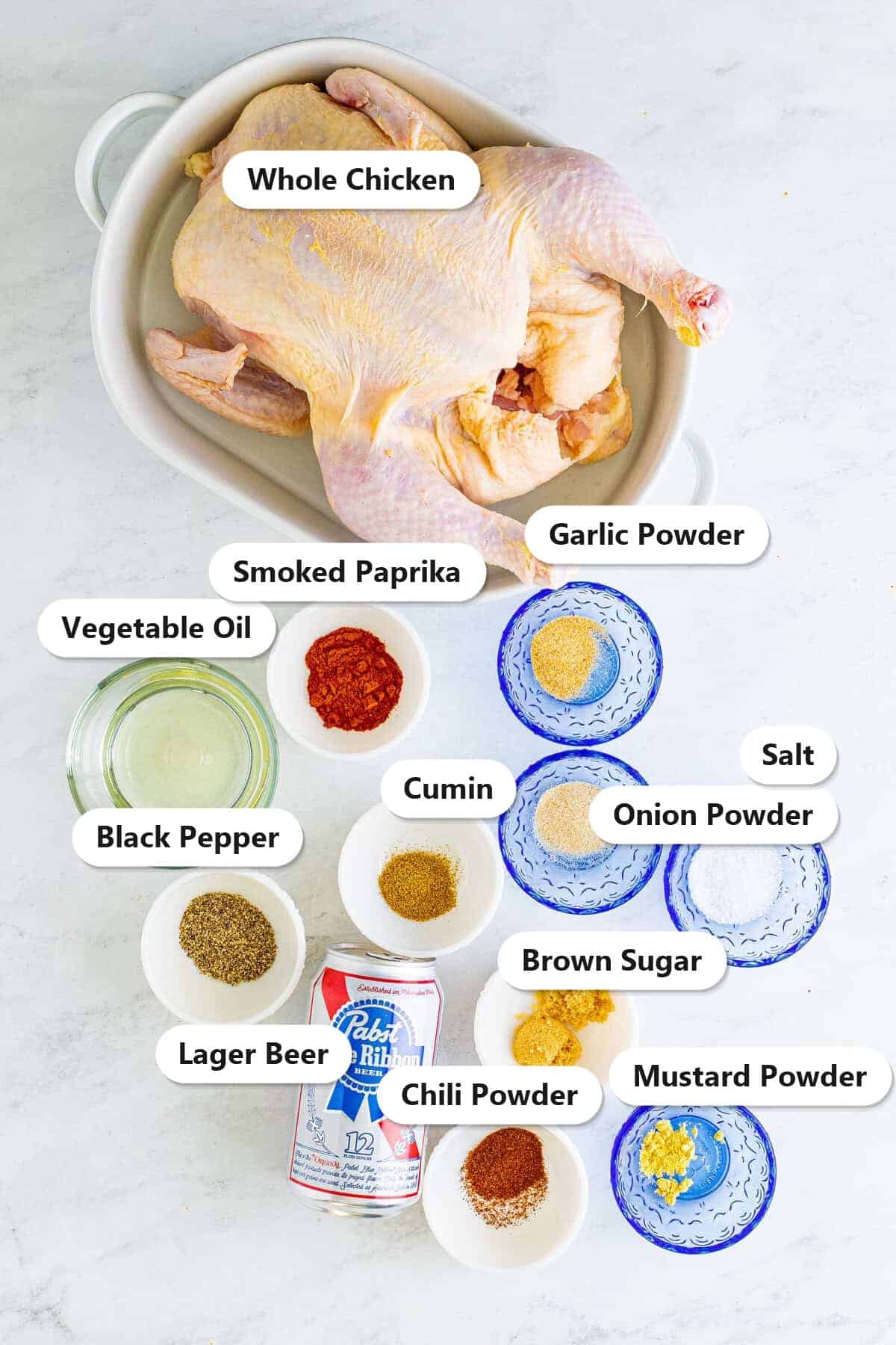Labeled ingredients for beer can chicken including a whole chicken, vegetable oil, smoked paprika, brown sugar, garlic powder, onion powder, cumin, chili powder, mustard powder, salt, black pepper, and a can of lager beer.