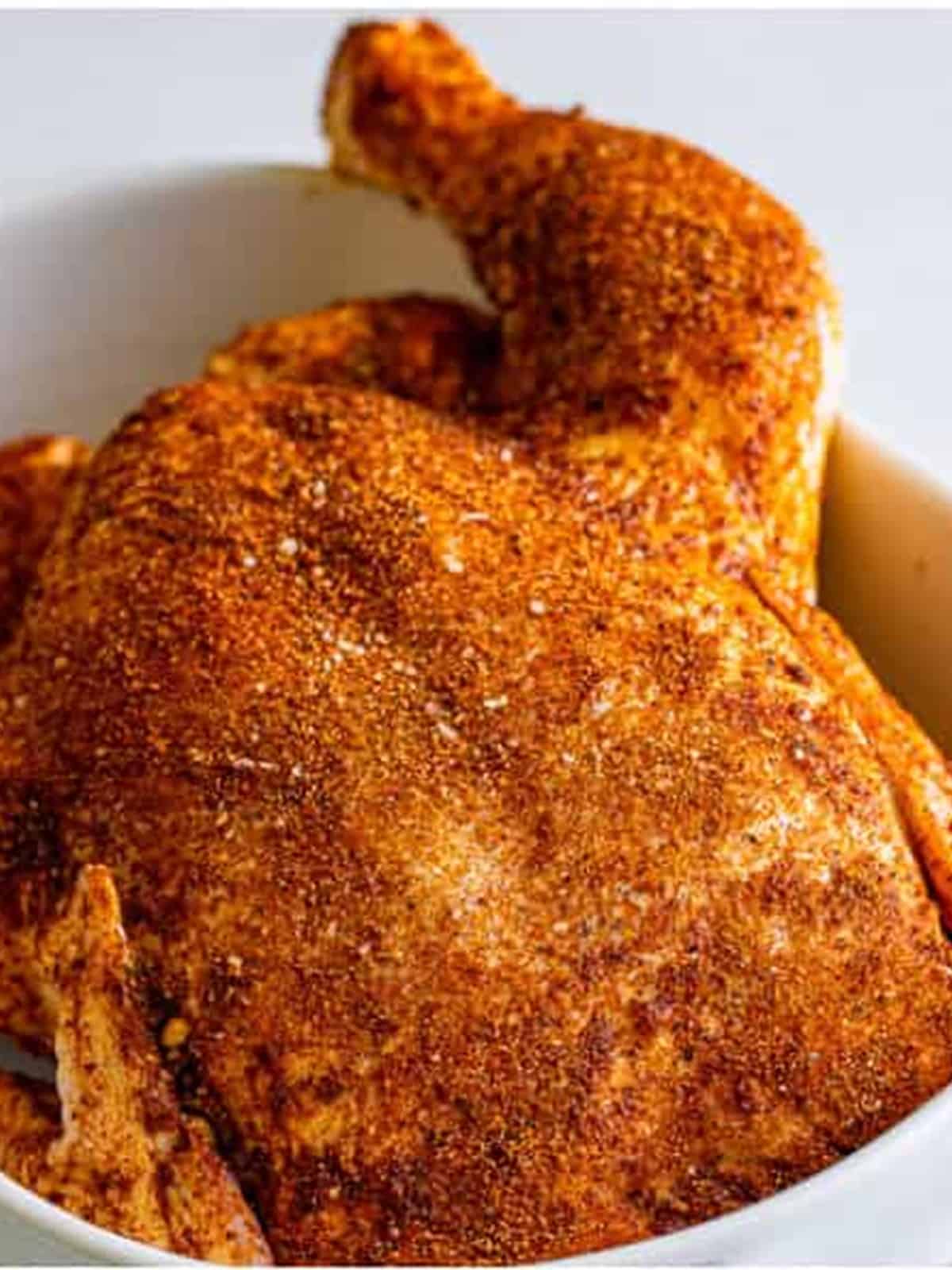 A whole raw chicken rubbed all over with smoked paprika spice rub in a white baking dish.