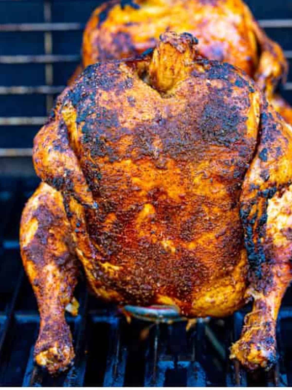 A whole chicken standing upright on a beer can on the grill grates cooking over indirect heat.