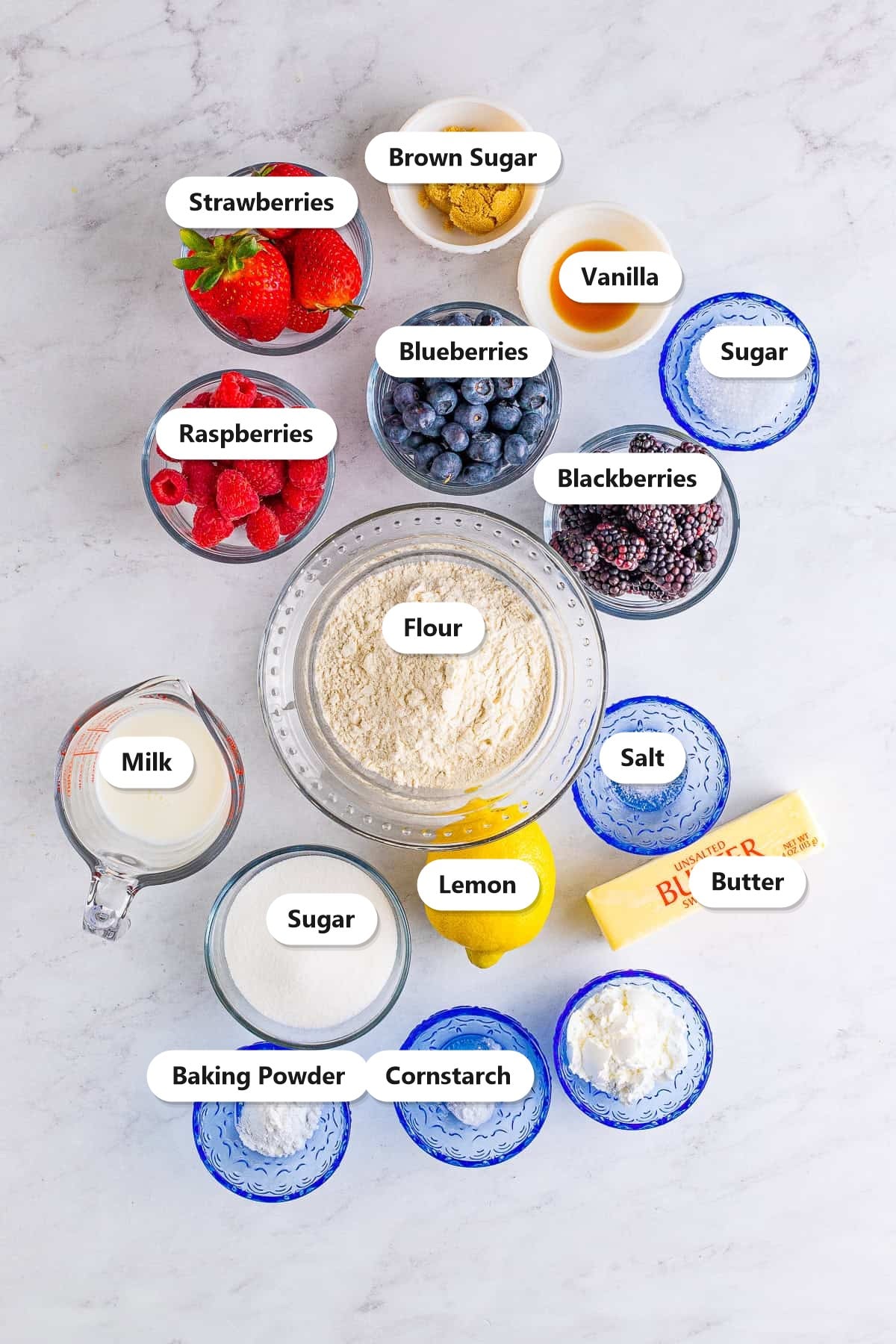 Labeled ingredients for berry cobbler including strawberries, blueberries, raspberries, blackberries, flour, sugar, butter, and lemon.