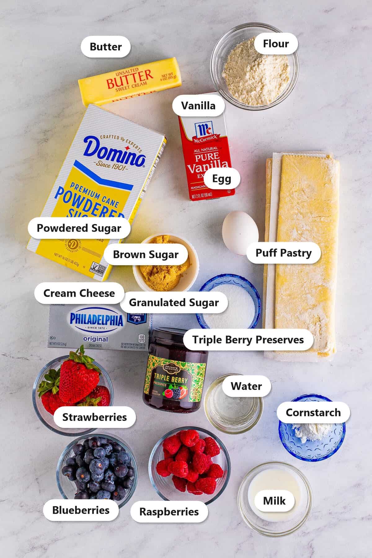 Labeled ingredients for berry danish including puff pastry, cream cheese, triple berry preserves, fresh blueberries, raspberries, and strawberries on a marble counter.