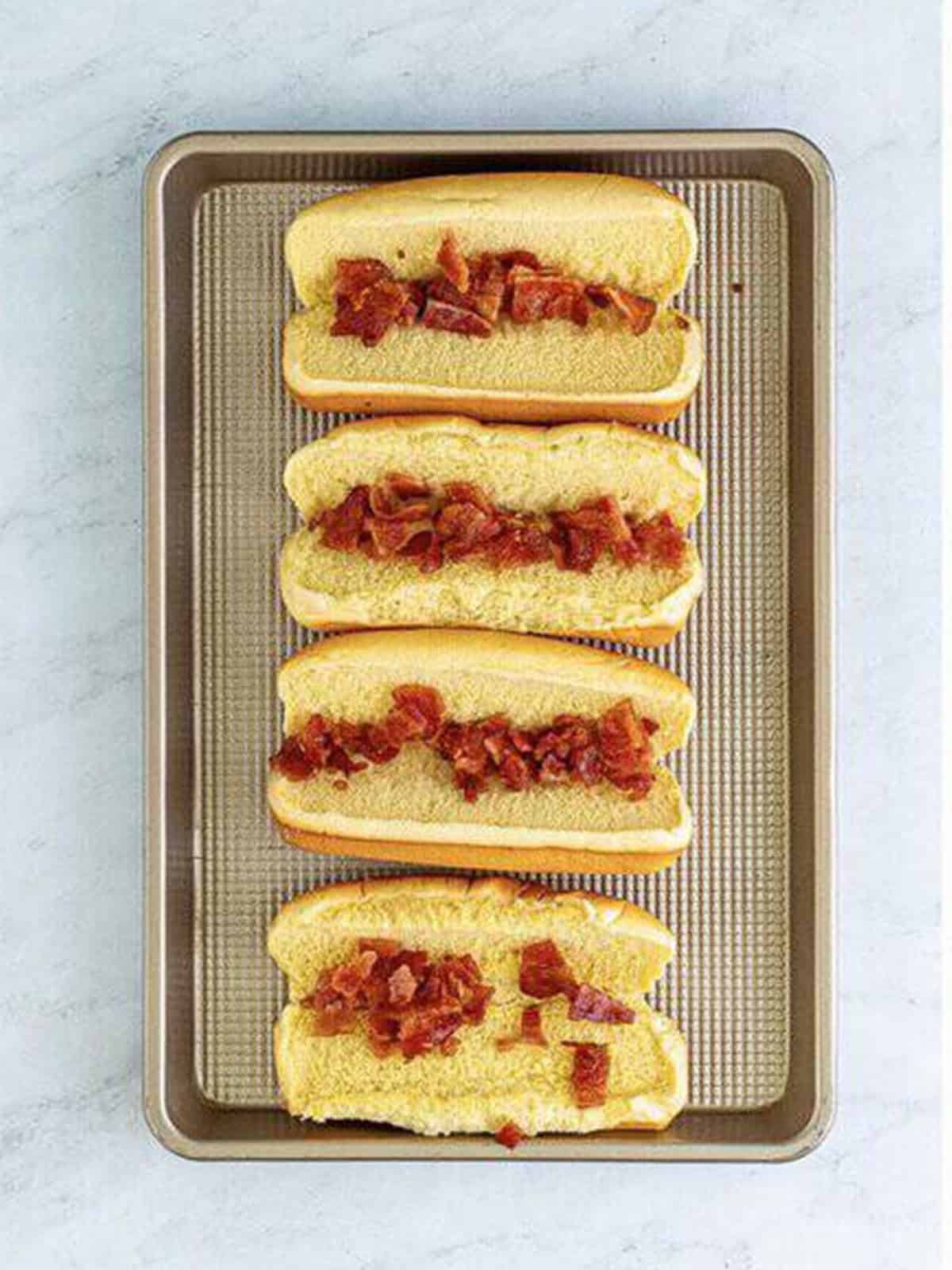 Crispy crumbled bacon added to the inside of hot dog buns on a sheet tray.