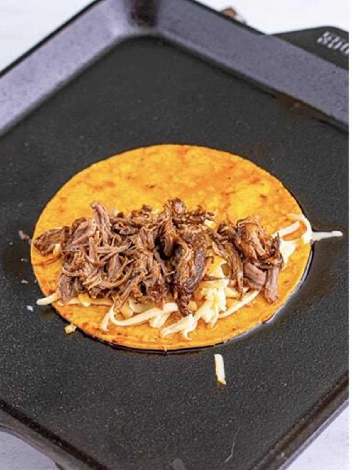 Corn tortilla filled with shredded birria beef and cheese on a griddle.