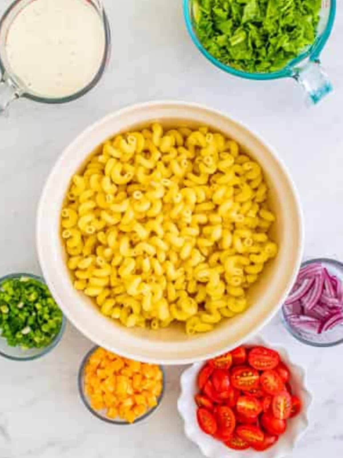 Cooked cavatappi pasta in a large bowl surrounded by prepped toppings for BLT Pasta Salad.