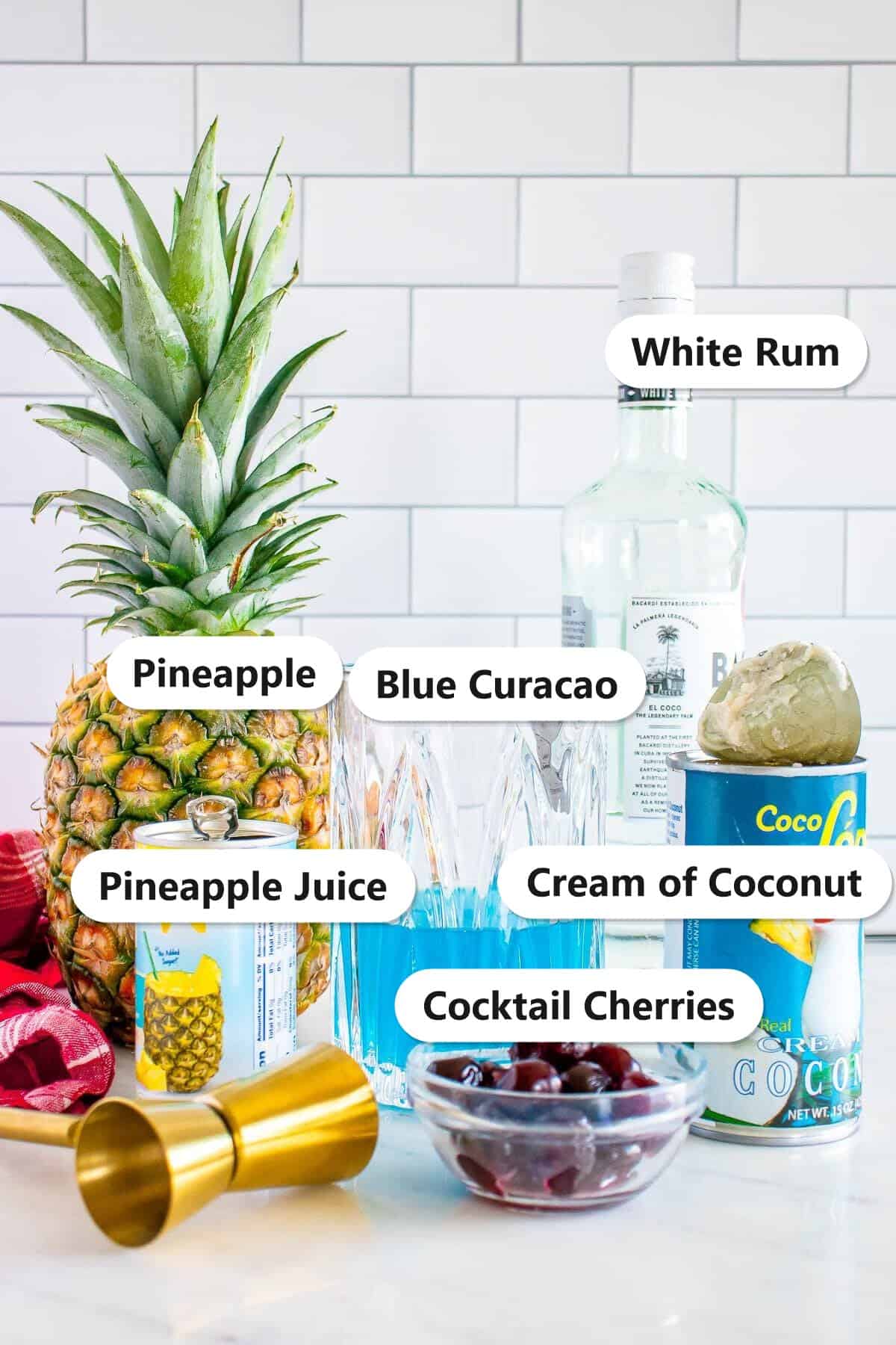 Labeled ingredients for a Blue Hawaiian Cocktail including white rum, blue curacao, pineapple juice, cream of coconut, fresh pineapple, and cocktail cherries.