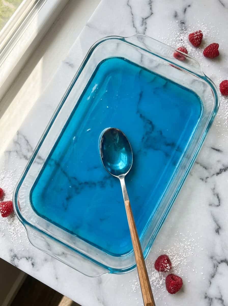 Bright blue raspberry Jello and soda mixture stirred in a glass baking dish before freezing into slush.