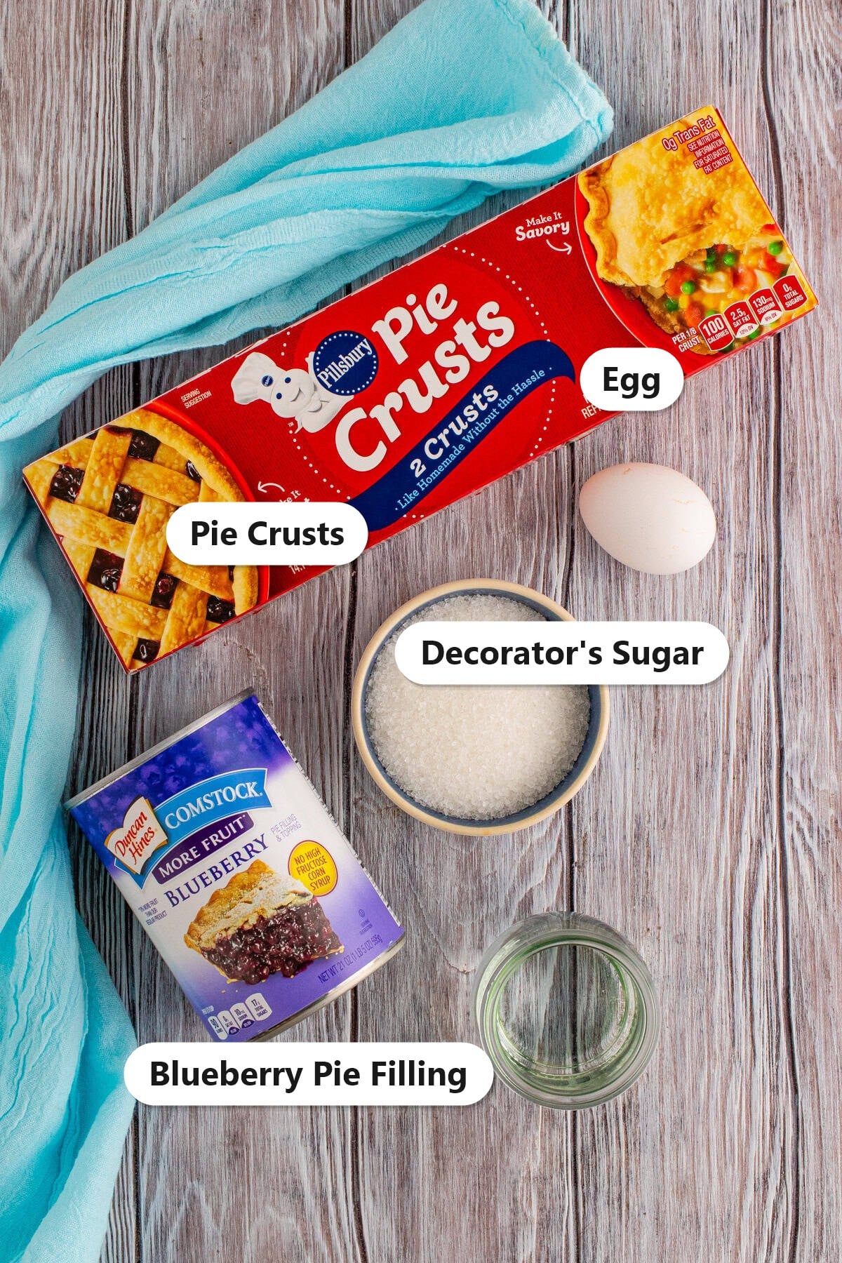 Labeled ingredients for blueberry hand pies including refrigerated pie crusts, blueberry pie filling, an egg, and decorators sugar.