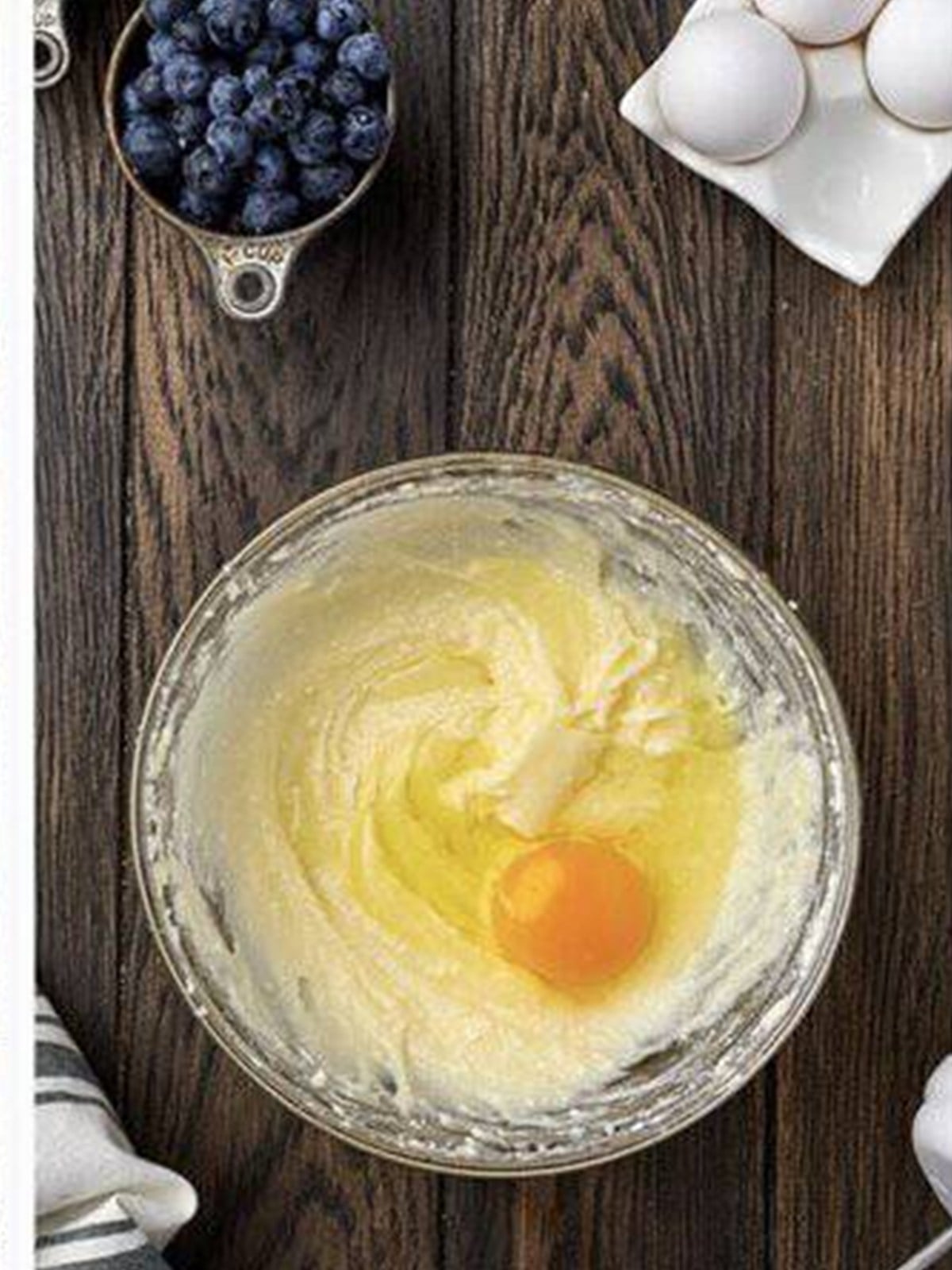 Blueberry muffin batter with an egg being mixed in a glass bowl.