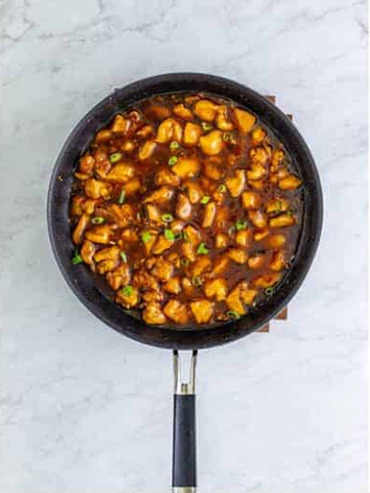 Finished bourbon chicken simmering in glossy sauce and garnished with scallions in a skillet.