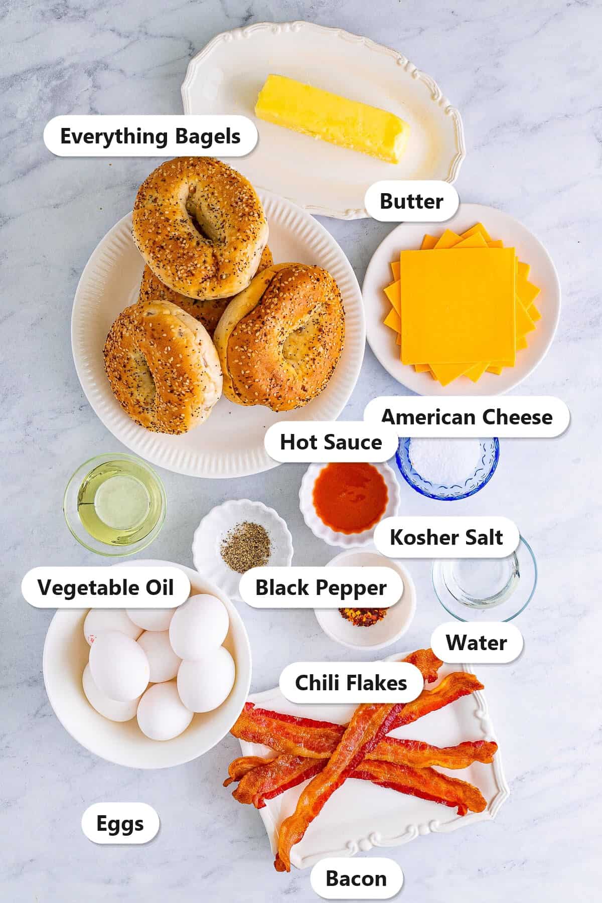 Labeled ingredients for a breakfast bagel sandwich including everything bagels, butter, American cheese, eggs, bacon, vegetable oil, hot sauce, kosher salt, black pepper, chili flakes, and water.