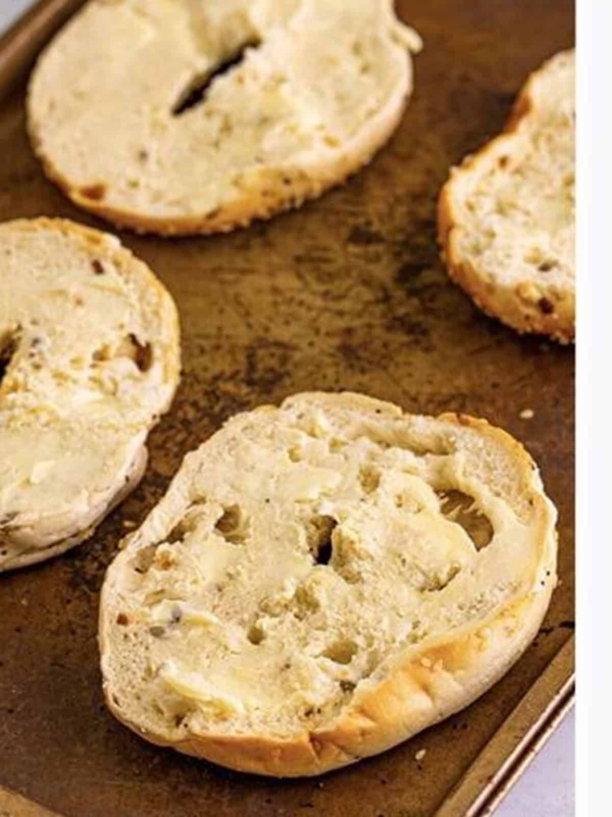 Everything bagel halves spread with butter cut side up on a sheet tray.