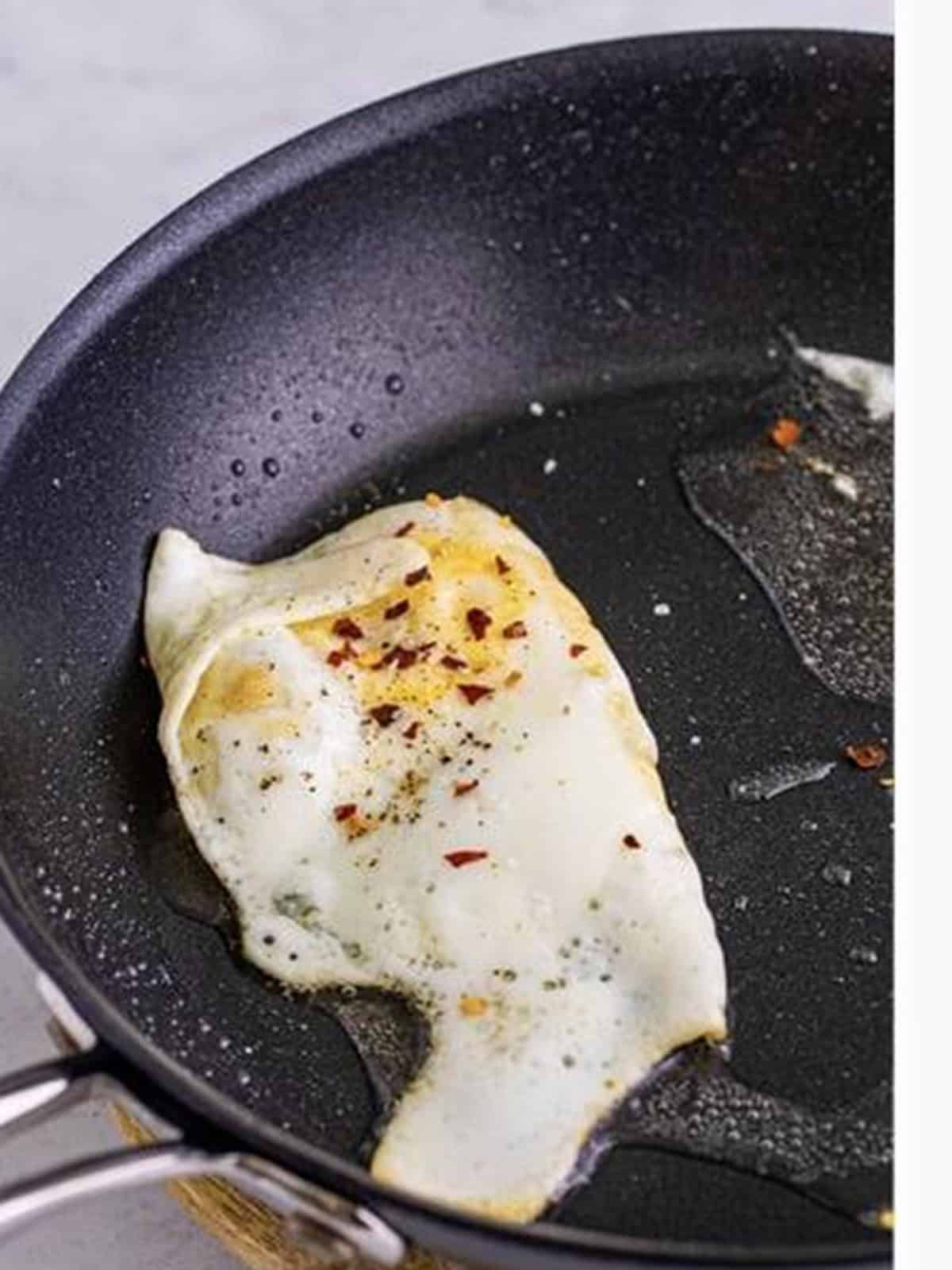 An egg frying in a nonstick skillet seasoned with salt, pepper, and chili flakes.
