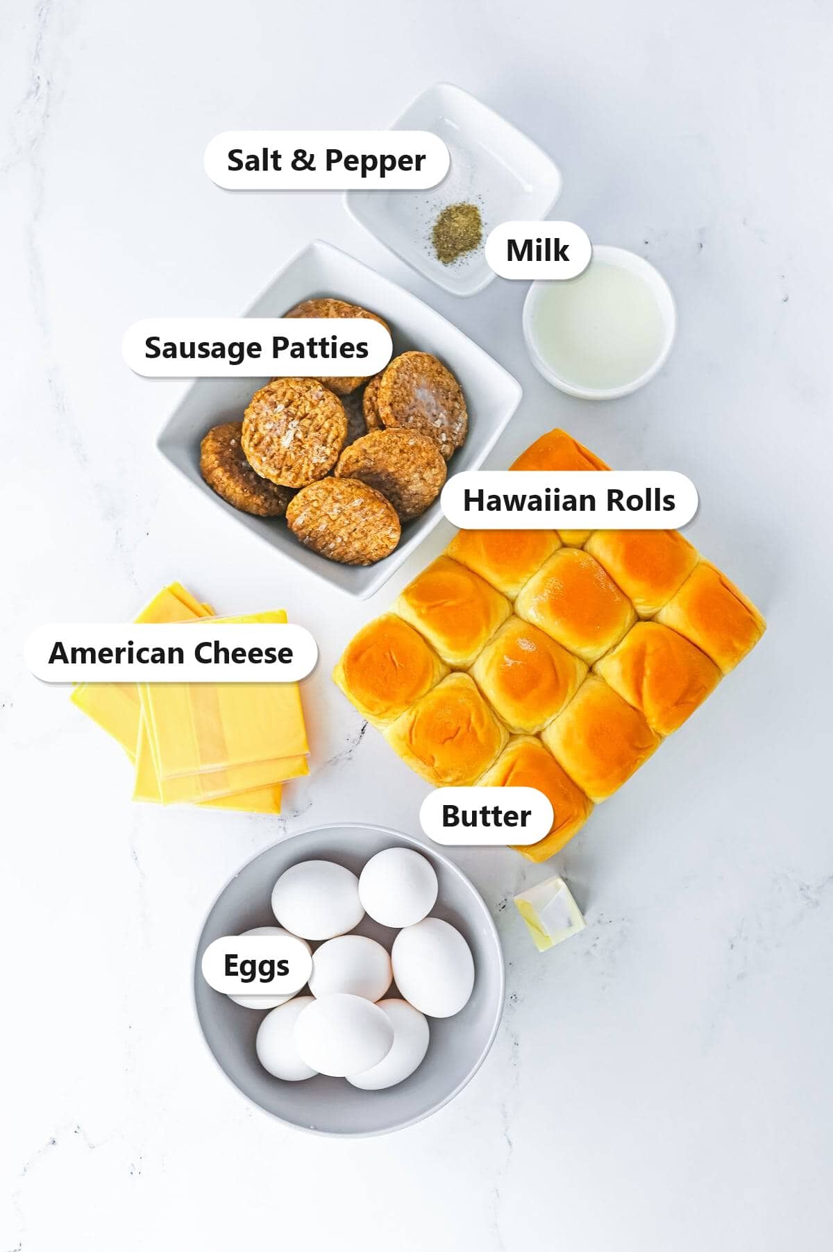 Labeled ingredients for sausage egg and cheese breakfast sliders including eggs, milk, salt and pepper, Hawaiian rolls, sausage patties, American cheese, and butter.