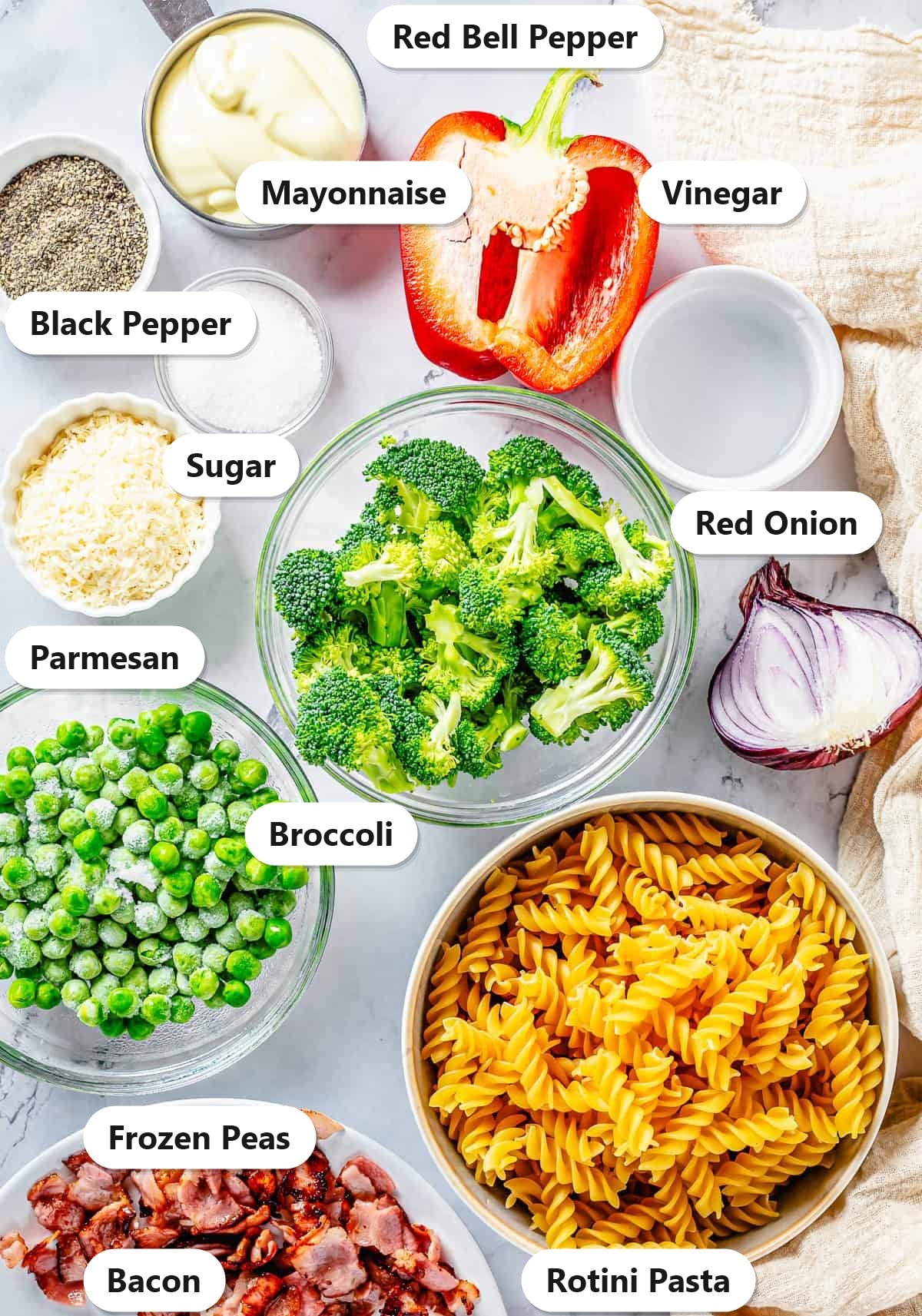 Labeled ingredients for broccoli pasta salad including rotini pasta, broccoli, bacon, frozen peas, red bell pepper, red onion, parmesan, mayonnaise, sugar, vinegar, and black pepper.