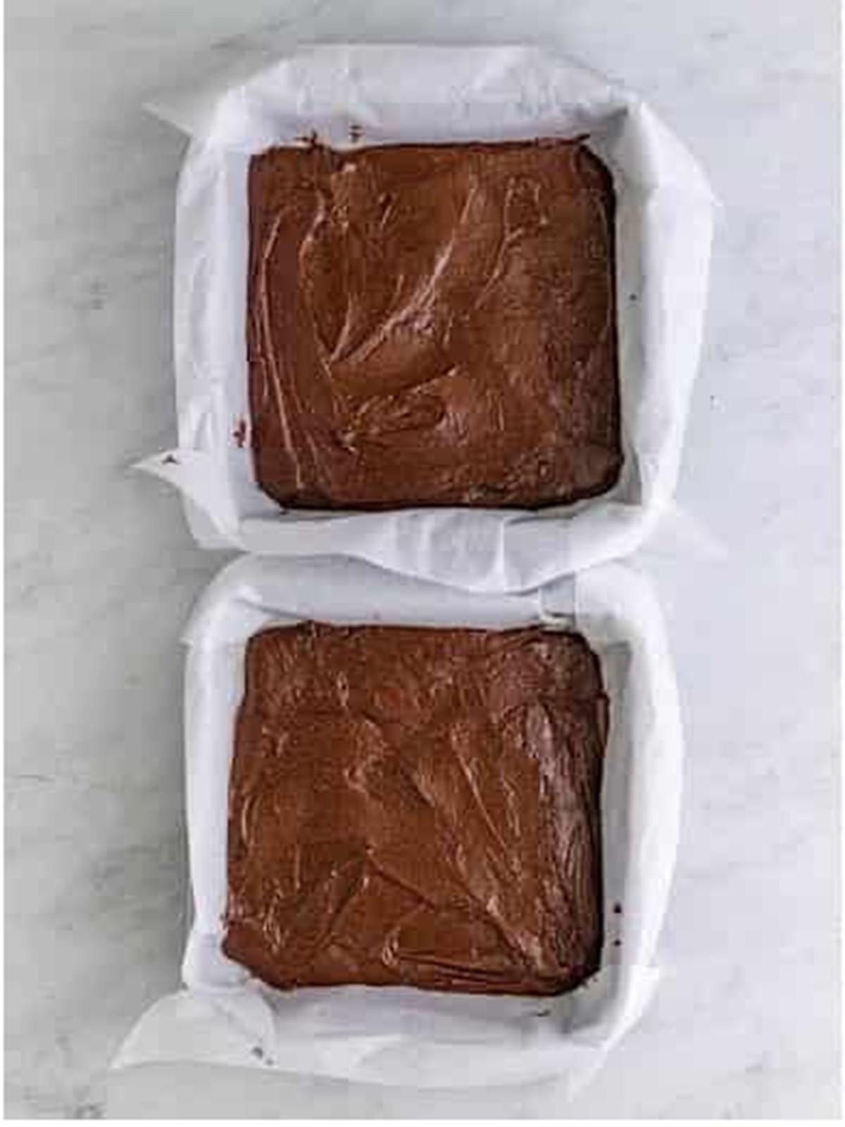Two baked brownie layers cooling in parchment lined square pans.