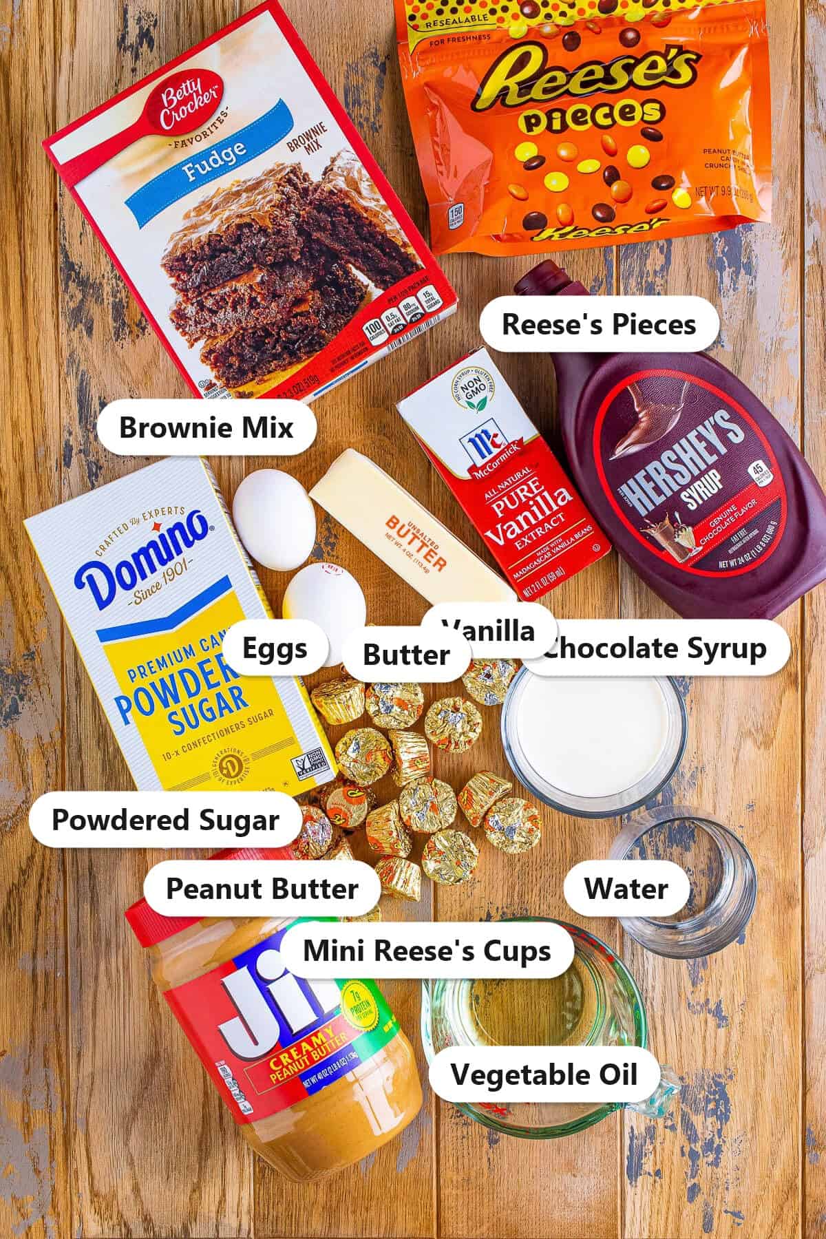 Labeled ingredients for brownie pizza including brownie mix, eggs, vegetable oil, water, peanut butter, butter, powdered sugar, vanilla, mini peanut butter cups, candy pieces, and chocolate syrup.