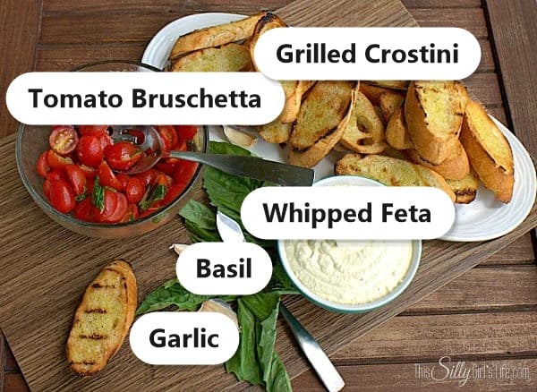Ingredients for bruschetta with whipped feta including marinated tomato bruschetta, grilled crostini, whipped feta, basil, and garlic.